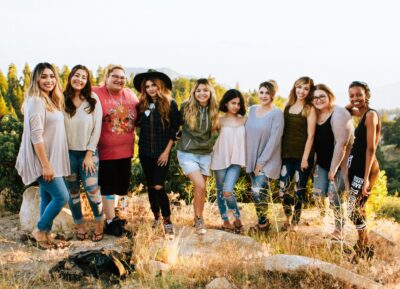 Eine Gruppe von zehn Frauen steht auf einem sonnigen Hügel inmitten einer natürlichen Landschaft. Sie lächeln fröhlich in die Kamera, umgeben von Büschen und Bäumen im Hintergrund.