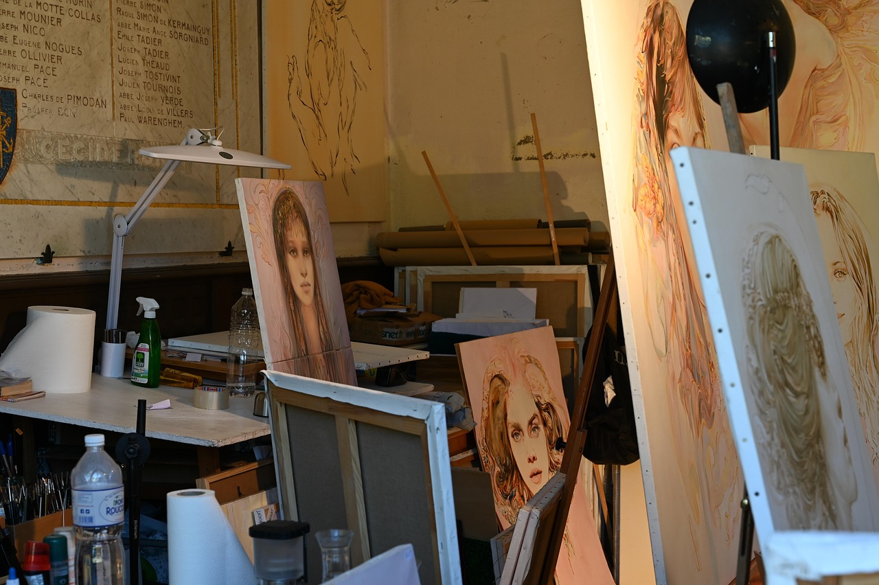 Künstleratelier mit mehreren Porträtzeichnungen oder -gemälden in warmen, pastelligen Tönen. Die Kunstwerke zeigen Frauenporträts in einem romantischen, skizzenhaften Stil. Im Vordergrund sind typische Ateliermaterialien zu sehen: eine Schreibtischlampe, Wasserflaschen, Malutensilien und leere Leinwände. An der Wand im Hintergrund ist eine Liste mit französischen Namen zu erkennen. Das Atelier wirkt authentisch und in Benutzung, mit einer Mischung aus Arbeitsplatz und künstlerischer Unordnung.