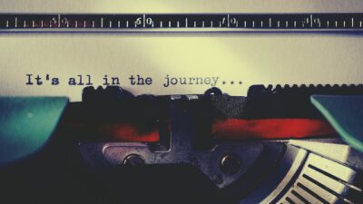 Nahaufnahme einer Schreibmaschine mit einem eingelegten Blatt Papier. Auf dem Papier steht der getippte Satz: ›It's all in the journey...‹. Die Szene ist in warmen, nostalgischen Farben gehalten.
