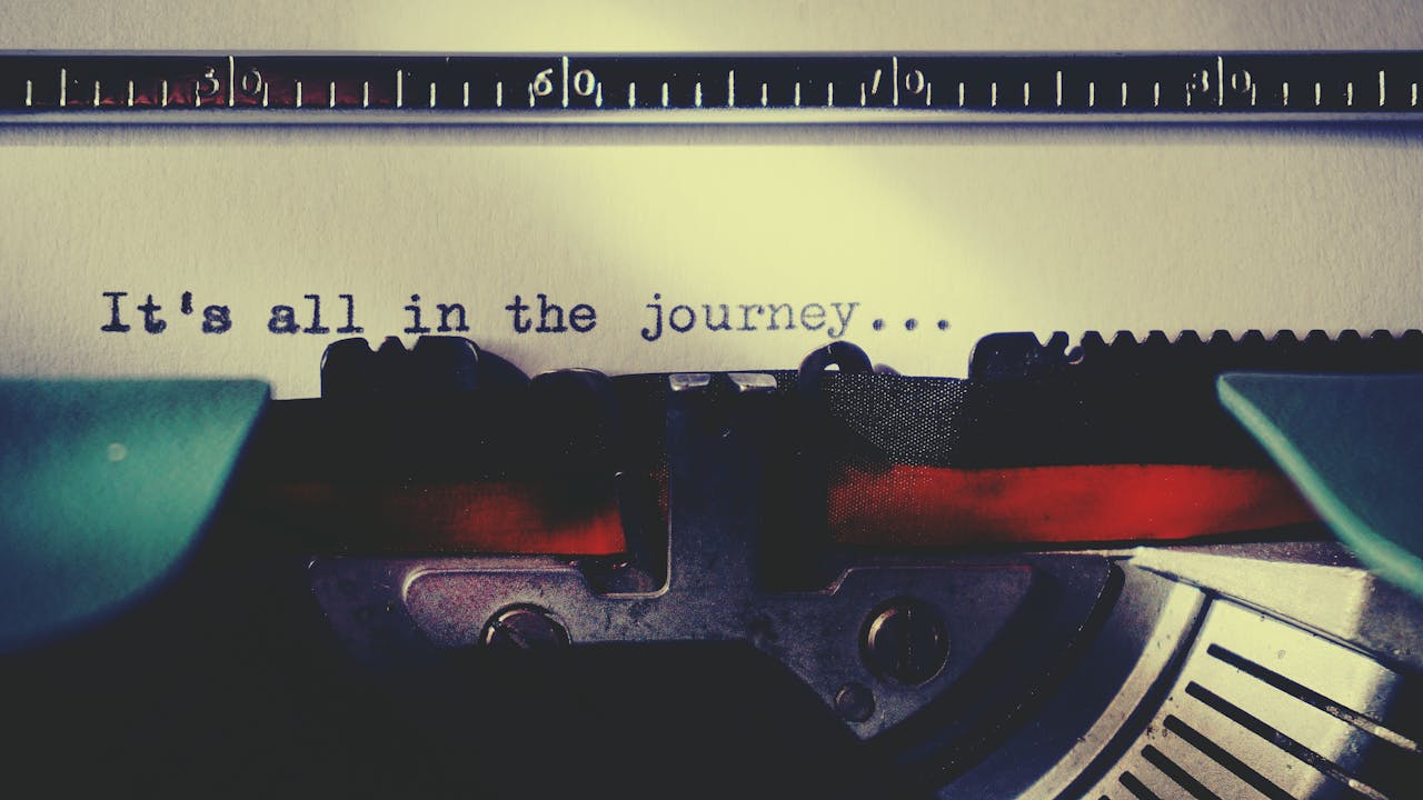 Nahaufnahme einer Schreibmaschine mit einem eingelegten Blatt Papier. Auf dem Papier steht der getippte Satz: ›It's all in the journey...‹. Die Szene ist in warmen, nostalgischen Farben gehalten.