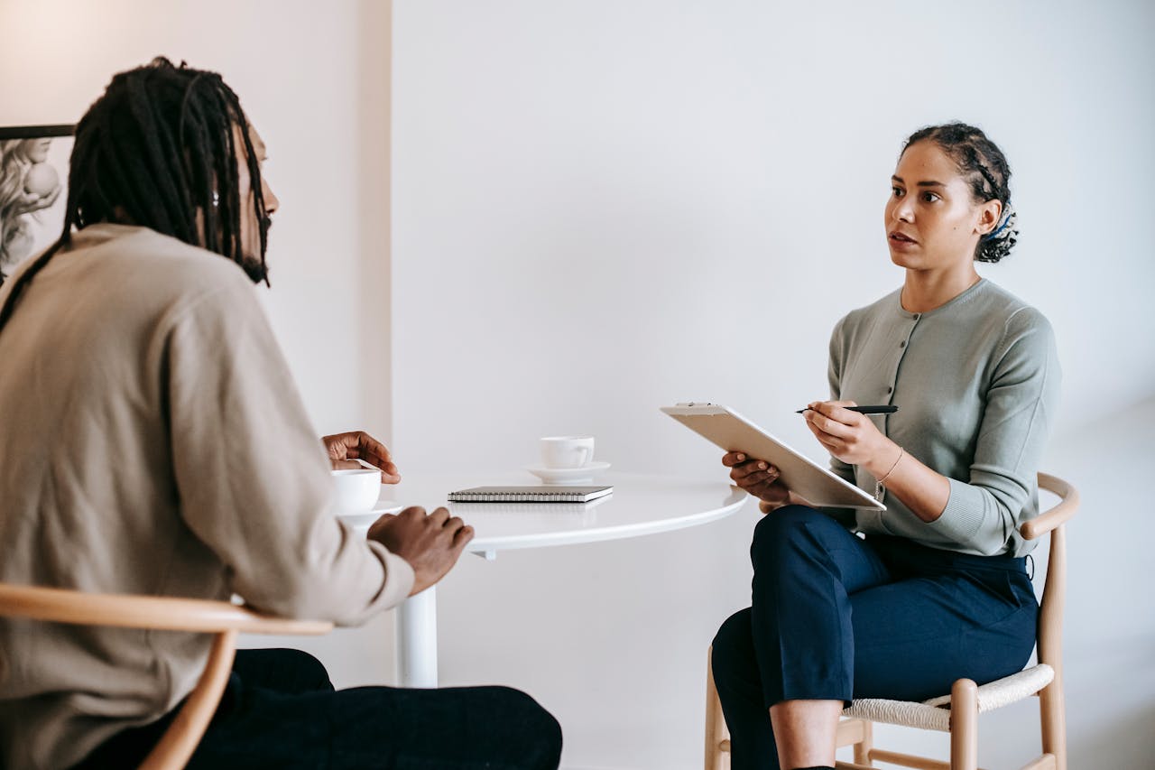 Eine Frau mit dunklen, geflochtenen Haaren sitzt an einem runden Tisch und führt ein Gespräch mit einem Mann mit Dreadlocks. Sie hält ein Klemmbrett und einen Stift in der Hand, während er eine Tasse Kaffee vor sich hat. Beide wirken konzentriert. Im Hintergrund ist eine weiße Wand mit einem gerahmten Bild zu sehen.