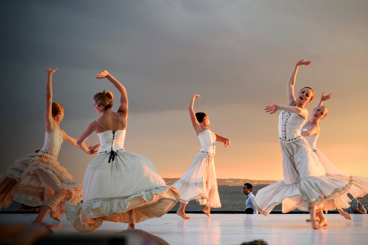 Eine Gruppe von Tänzerinnen in weißen, fließenden Kleidern tanzt anmutig auf einer Open-Air-Bühne vor einer malerischen Kulisse mit Sonnenuntergang und Meer im Hintergrund. Ihre Bewegungen wirken elegant und synchron.