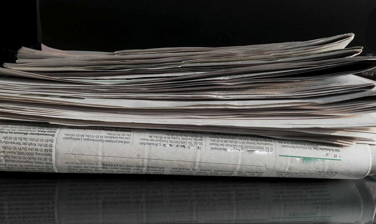 Ein Stapel alter, gefalteter Zeitungen liegt auf einer reflektierenden Oberfläche. Die Blätter sind leicht zerknittert und übereinander gestapelt. Der Hintergrund ist dunkel, wodurch der Zeitungsstapel im Vordergrund hervorsticht.