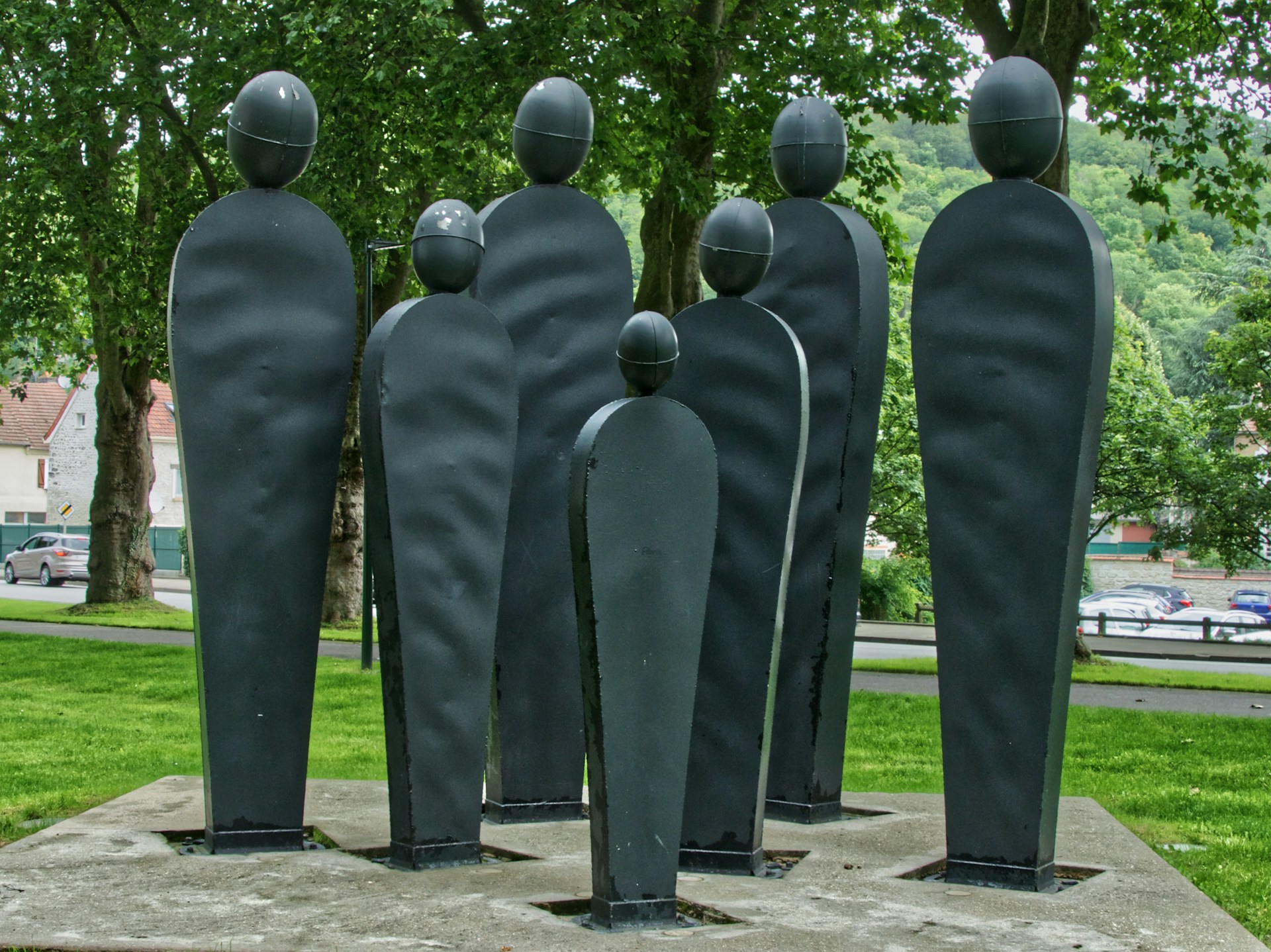 Moderne Skulpturengruppe im Freien, bestehend aus acht abstrakten, stilisierten menschlichen Figuren aus dunklem Metall mit ovalen Köpfen. Die Figuren stehen auf einem Betonfundament in einem grünen Park mit Bäumen im Hintergrund.
