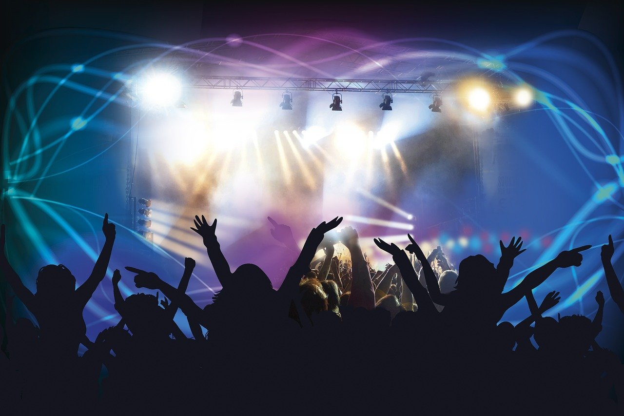 Menschenmenge bei einem Live-Konzert mit erhobenen Armen vor einer hell beleuchteten Bühne, auf der farbige Lichtstrahlen und Scheinwerfer eine dynamische Atmosphäre erzeugen. Im Vordergrund sind die Zuschauer:innen als dunkle Silhouetten zu sehen.