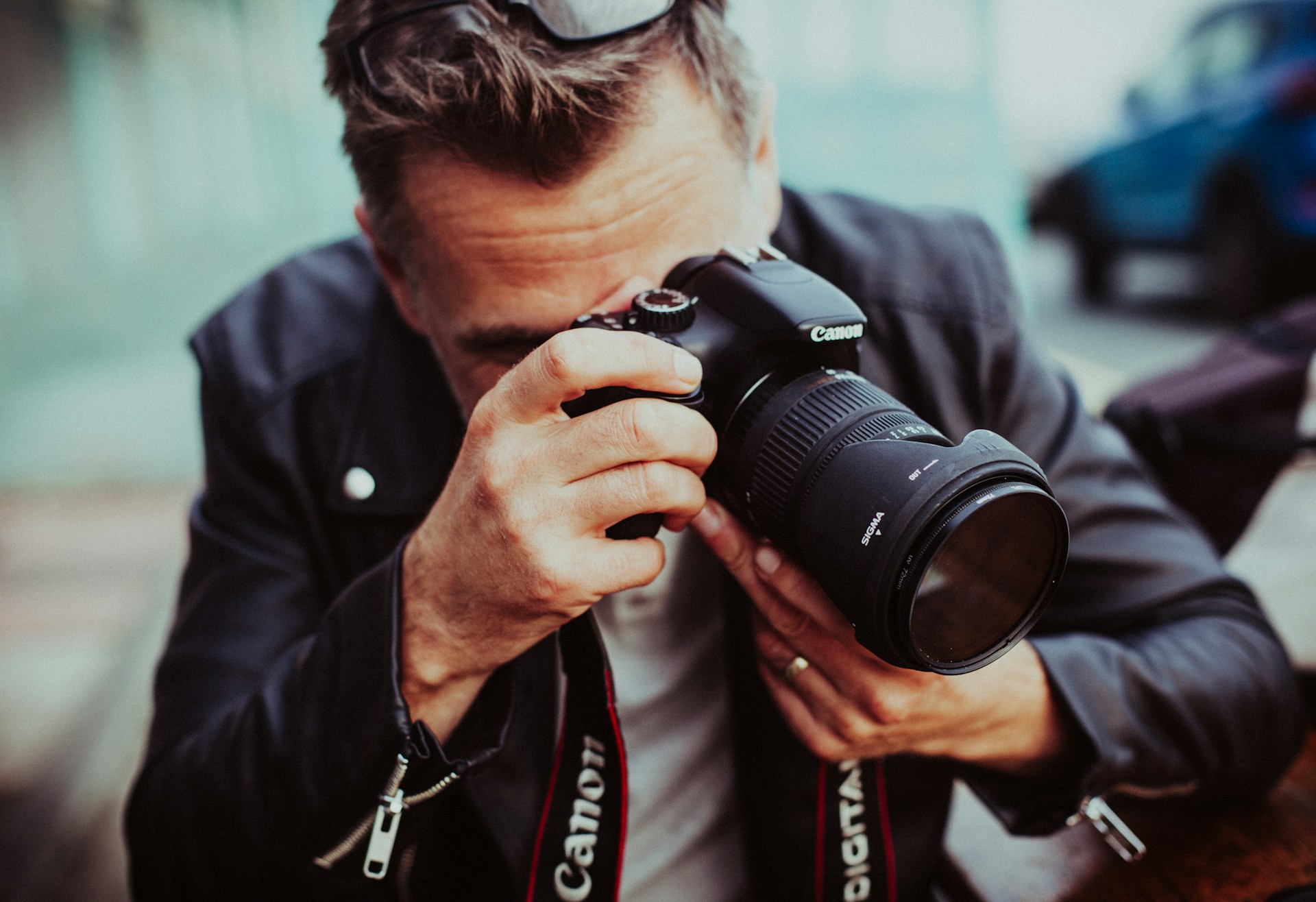 Ein Mann in Lederjacke hält eine Canon-Spiegelreflexkamera mit Sigma-Objektiv vor sein Gesicht und macht ein Foto; er sitzt im Freien und trägt eine Sonnenbrille auf dem Kopf.