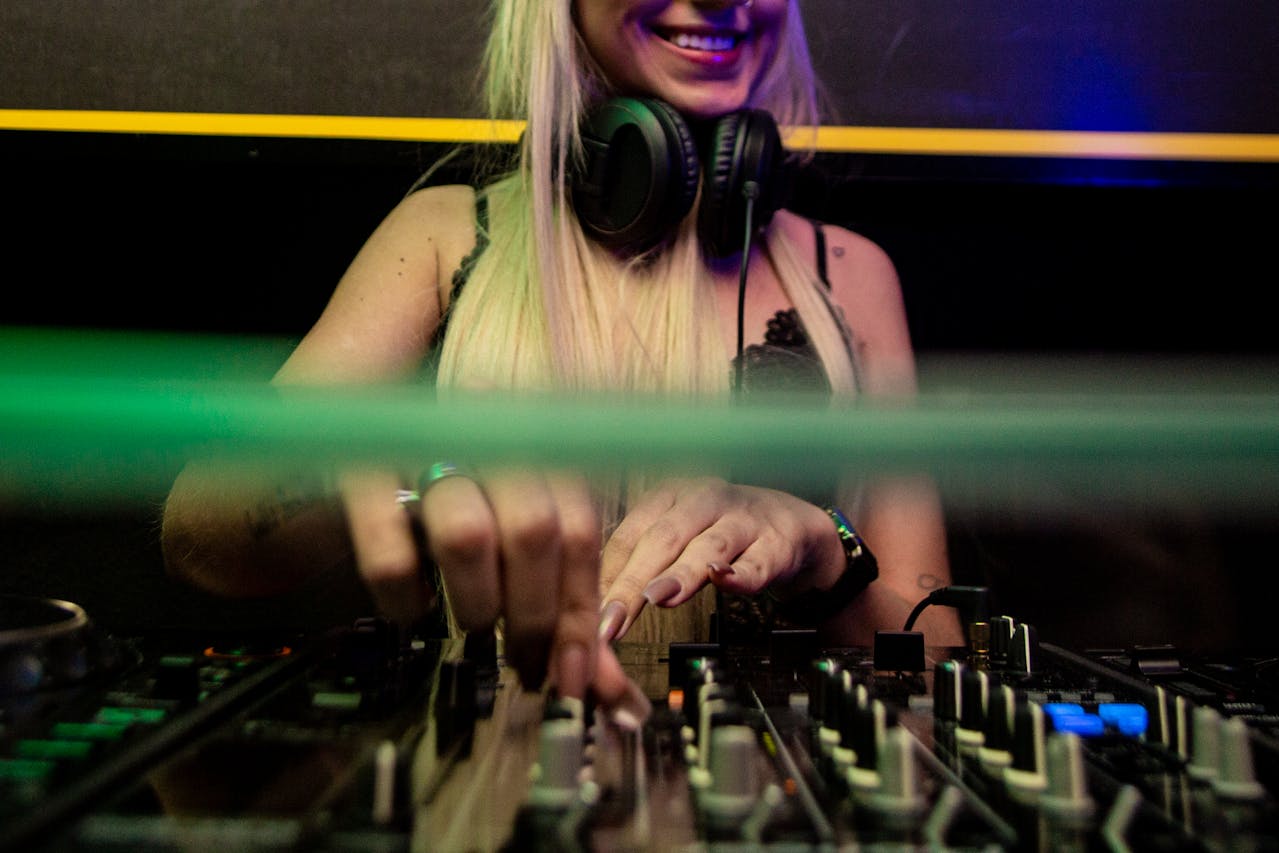 Eine junge DJane mit langen blonden Haaren und Kopfhörern um den Hals steht an einem Mischpult und legt auf. Sie lächelt, während sie konzentriert an den Reglern arbeitet. Das Foto zeigt eine Nahaufnahme ihrer Hände und des Mischpults in einem clubähnlichen Setting mit farbigen Lichtern.