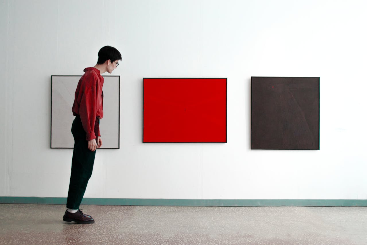 Eine Person in rotem Hemd und schwarzer Hose betrachtet in einer Galerie drei abstrakte, quadratische Gemälde an einer weißen Wand – eines weiß, eines rot und eines dunkelbraun.