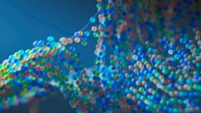 Bunte, unscharf dargestellte Molekülstruktur auf blauem Hintergrund; zahlreiche kleine, farbige Kugeln sind durch dünne Verbindungen zu einem komplexen, netzartigen Muster verbunden, das an eine DNA-Doppelhelix erinnert.