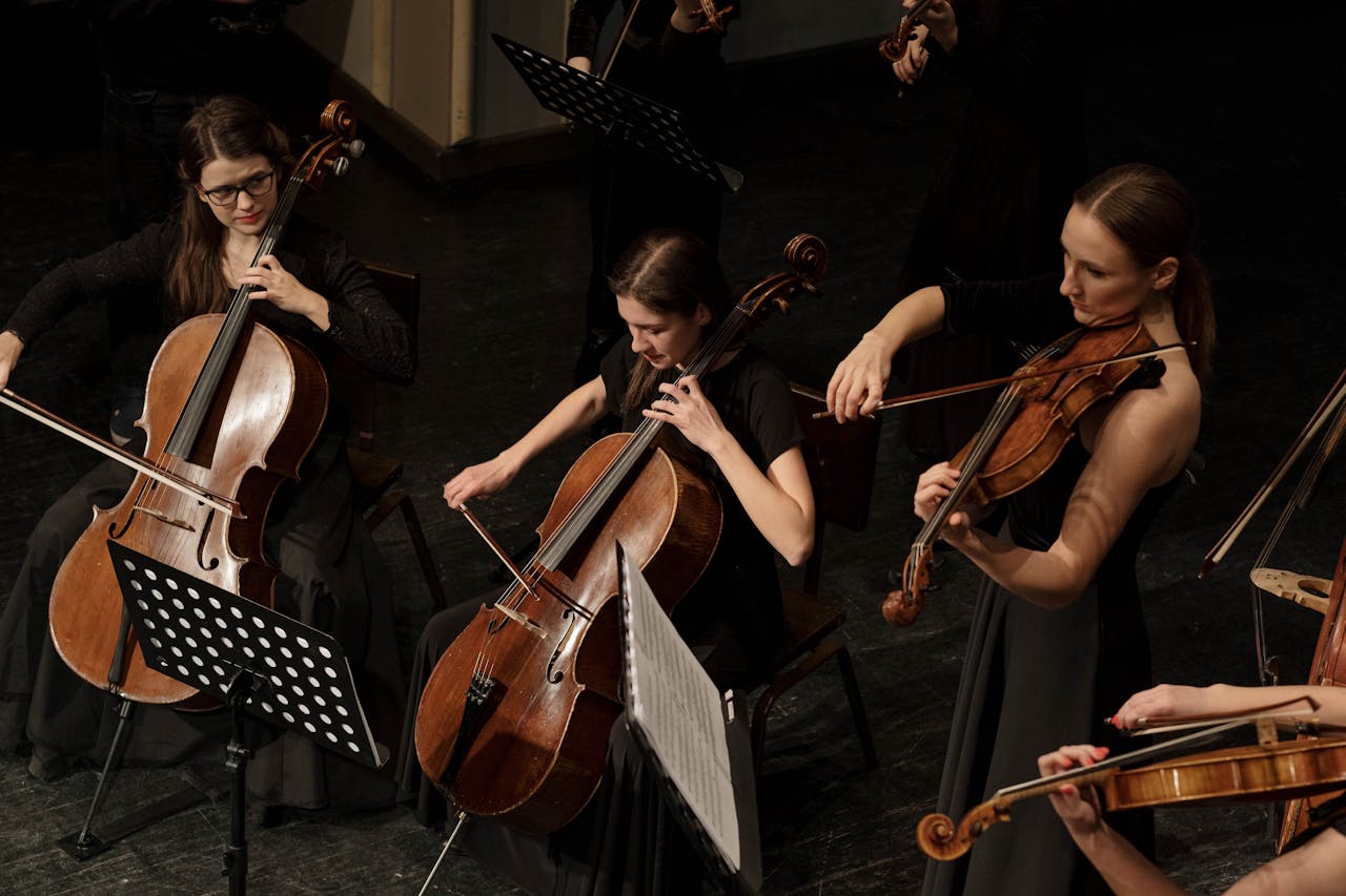 Mehrere Musikerinnen in schwarzer Konzertkleidung spielen Streichinstrumente wie Cello und Geige in einem klassischen Orchester auf einer Bühne.