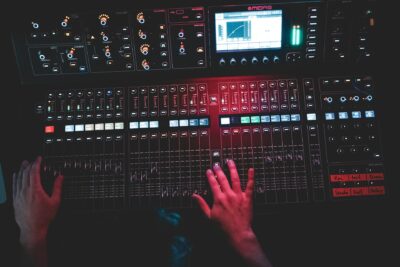 Ein Mensch bedient ein professionelles Audiomischpult mit zahlreichen Schiebereglern, Tasten, Drehknöpfen und einem digitalen Display. Die Szene ist dunkel und in rotes Licht getaucht, wodurch eine atmosphärische Studio- oder Live-Umgebung entsteht.