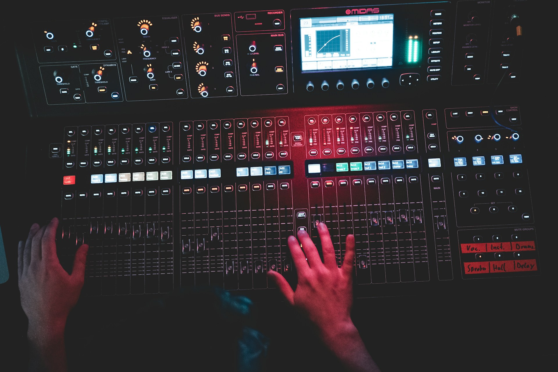 Ein Mensch bedient ein professionelles Audiomischpult mit zahlreichen Schiebereglern, Tasten, Drehknöpfen und einem digitalen Display. Die Szene ist dunkel und in rotes Licht getaucht, wodurch eine atmosphärische Studio- oder Live-Umgebung entsteht.