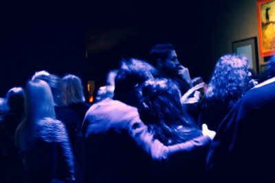 Eine Gruppe von Menschen steht dicht gedrängt in einem dunklen Raum mit schwacher, bläulicher Beleuchtung. Einige Personen scheinen sich zu unterhalten, während ein Paar sich innig umarmt. Im Hintergrund sind verschwommene Umrisse von Bildern an der Wand zu erkennen, eines davon zeigt eine Figur im Sonnenlicht. Die Szene wirkt wie auf einer Party oder bei einer Kunstausstellung, leicht verschwommen durch Bewegung.