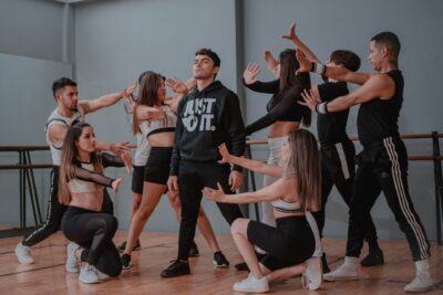 Eine Tanzgruppe aus acht jungen Erwachsenen führt eine choreografierte Pose in einem Tanzstudio auf. In der Mitte steht ein Mann mit schwarzem Hoodie, auf dem „JUST DO IT.“ steht, während die anderen ihn mit ausgestreckten Armen und dramatischen Gesten umkreisen. Die Szene wirkt dynamisch und ausdrucksstark, typisch für eine moderne Tanzaufführung oder Probe.
