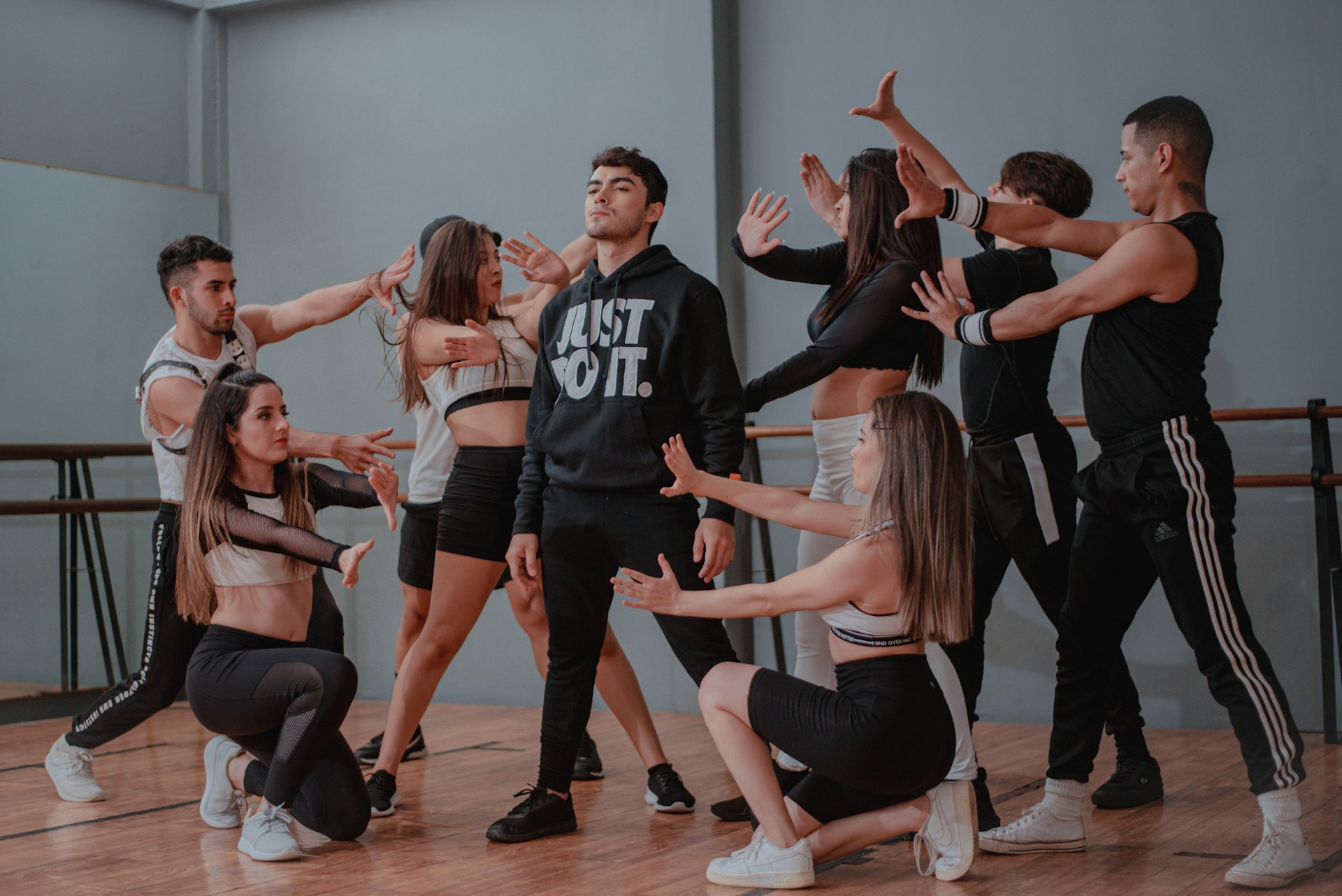 Eine Tanzgruppe aus acht jungen Erwachsenen führt eine choreografierte Pose in einem Tanzstudio auf. In der Mitte steht ein Mann mit schwarzem Hoodie, auf dem „JUST DO IT.“ steht, während die anderen ihn mit ausgestreckten Armen und dramatischen Gesten umkreisen. Die Szene wirkt dynamisch und ausdrucksstark, typisch für eine moderne Tanzaufführung oder Probe.