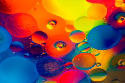 Bunte Flüssigkeitstropfen in leuchtenden Farben wie Blau, Rot, Gelb, Orange und Pink schweben nebeneinander und übereinander in einer abstrakten Komposition. Die Formen erinnern an Öl- oder Wassertropfen unter einem Mikroskop, die Licht in regenbogenartigen Farben reflektieren. Das Bild wirkt lebendig und künstlerisch.