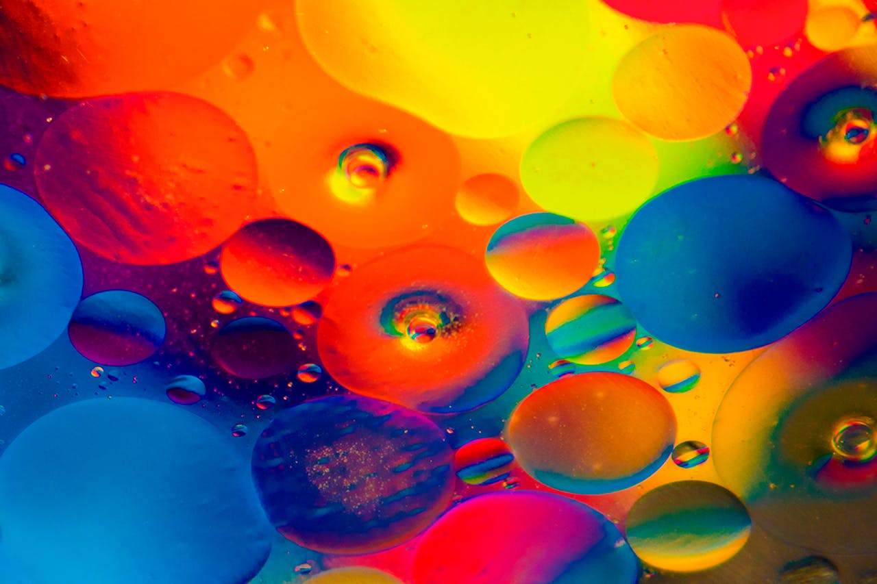 Bunte Flüssigkeitstropfen in leuchtenden Farben wie Blau, Rot, Gelb, Orange und Pink schweben nebeneinander und übereinander in einer abstrakten Komposition. Die Formen erinnern an Öl- oder Wassertropfen unter einem Mikroskop, die Licht in regenbogenartigen Farben reflektieren. Das Bild wirkt lebendig und künstlerisch.