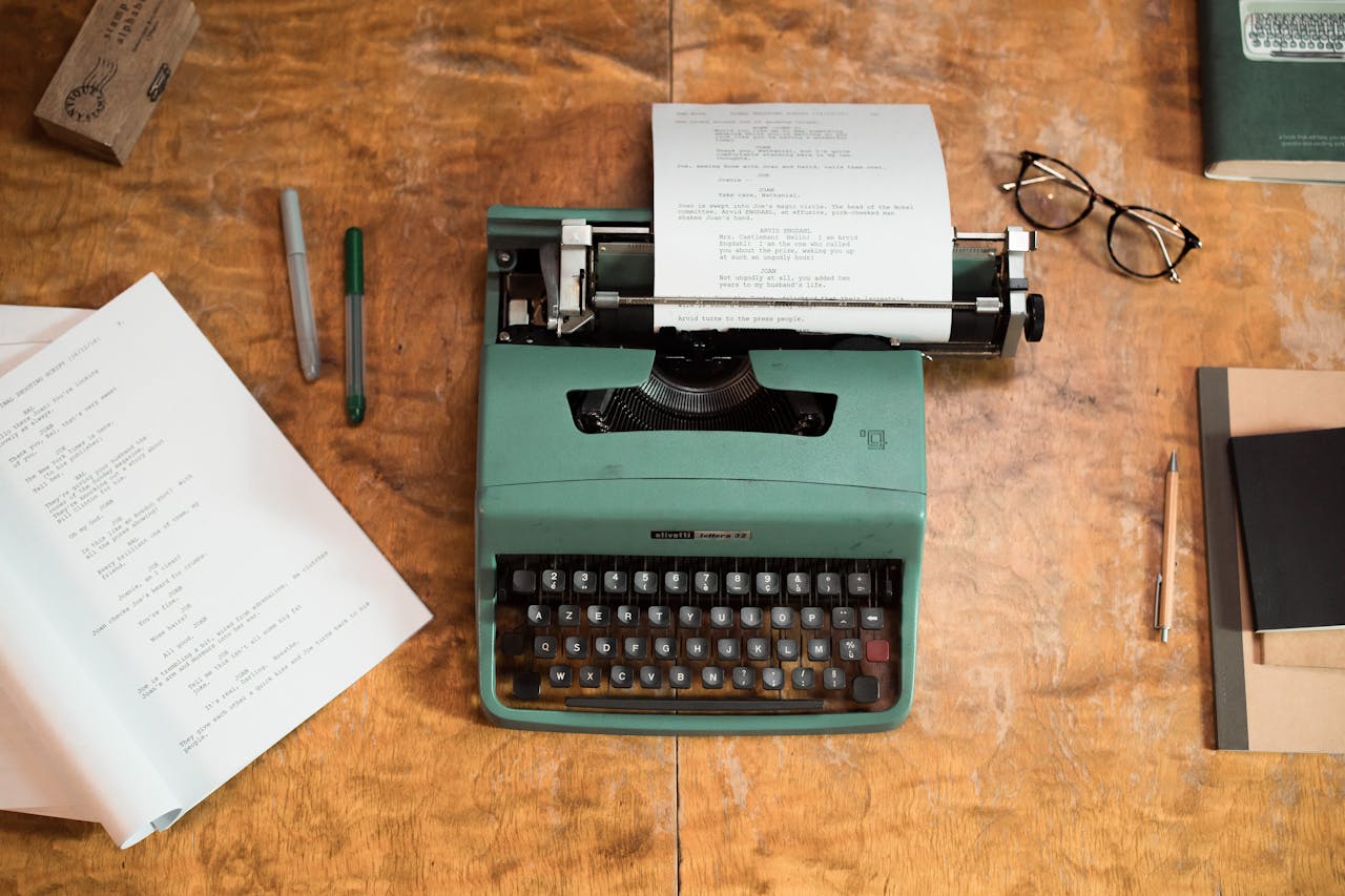 Eine grüne Schreibmaschine (Olivetti Lettera 32) steht auf einem hölzernen Schreibtisch. In der Maschine steckt ein getipptes Drehbuch oder Manuskript. Um die Schreibmaschine liegen zwei Stifte, ein aufgeklapptes Skript, ein Notizblock mit Stift, eine Brille und ein Buch. Die Szene wirkt nostalgisch und kreativ, als wäre jemand gerade beim Schreiben.