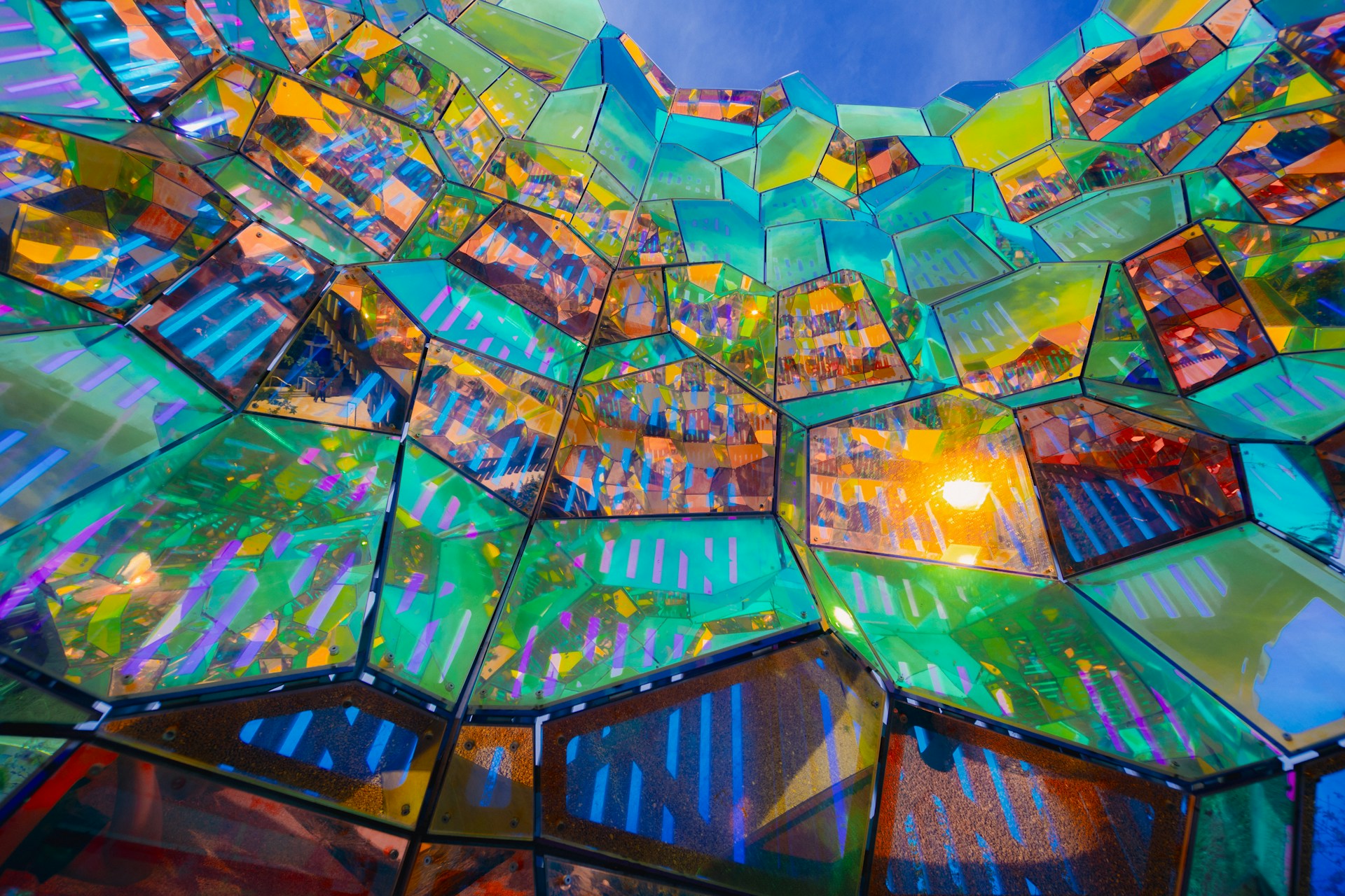 Bunte, facettenreiche Struktur aus transparenten, geometrischen Glaspaneelen in leuchtenden Farben wie Türkis, Gelb, Violett und Orange; das Licht bricht sich darin spektakulär und erzeugt lebendige Reflexionen und Muster vor einem blauen Himmel.