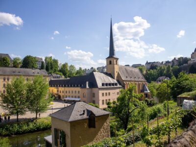 Historisches Gebäudeensemble mit einer großen Kirche und spitzem Turm in Luxemburg-Stadt, umgeben von Bäumen und alten Gemäuern an einem sonnigen Tag mit blauem Himmel und vereinzelten Wolken. Im Vordergrund ein kleiner Fluss und Gärten mit Weinreben.