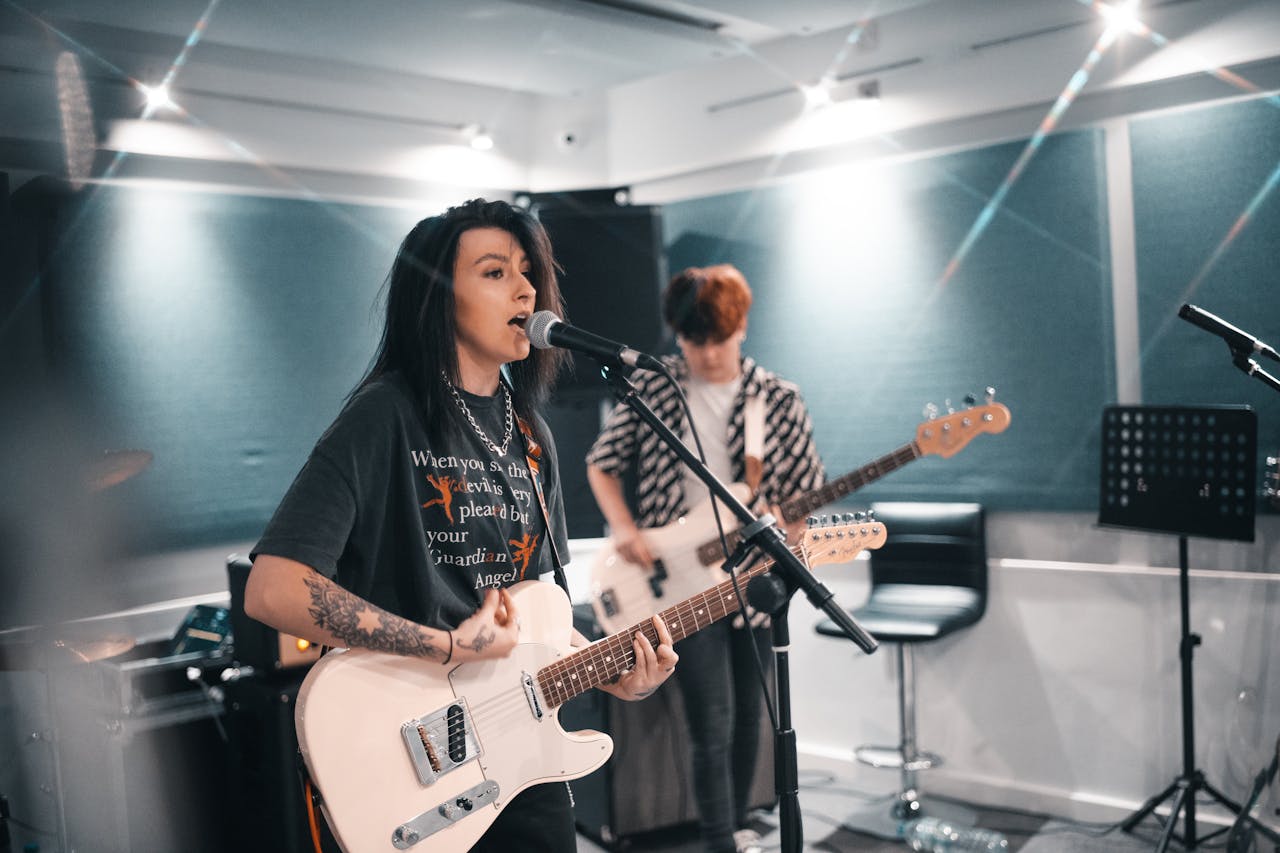 Zwei junge Musiker:innen in einem Proberaum. Im Vordergrund steht eine Person mit dunklen Haaren und Tätowierungen, die in ein Mikrofon singt und eine E-Gitarre spielt. Im Hintergrund spielt eine weitere Person Bassgitarre. Der Raum ist modern mit schallgedämpften Wänden ausgestattet, mehrere Mikrofone und Verstärker sind sichtbar.