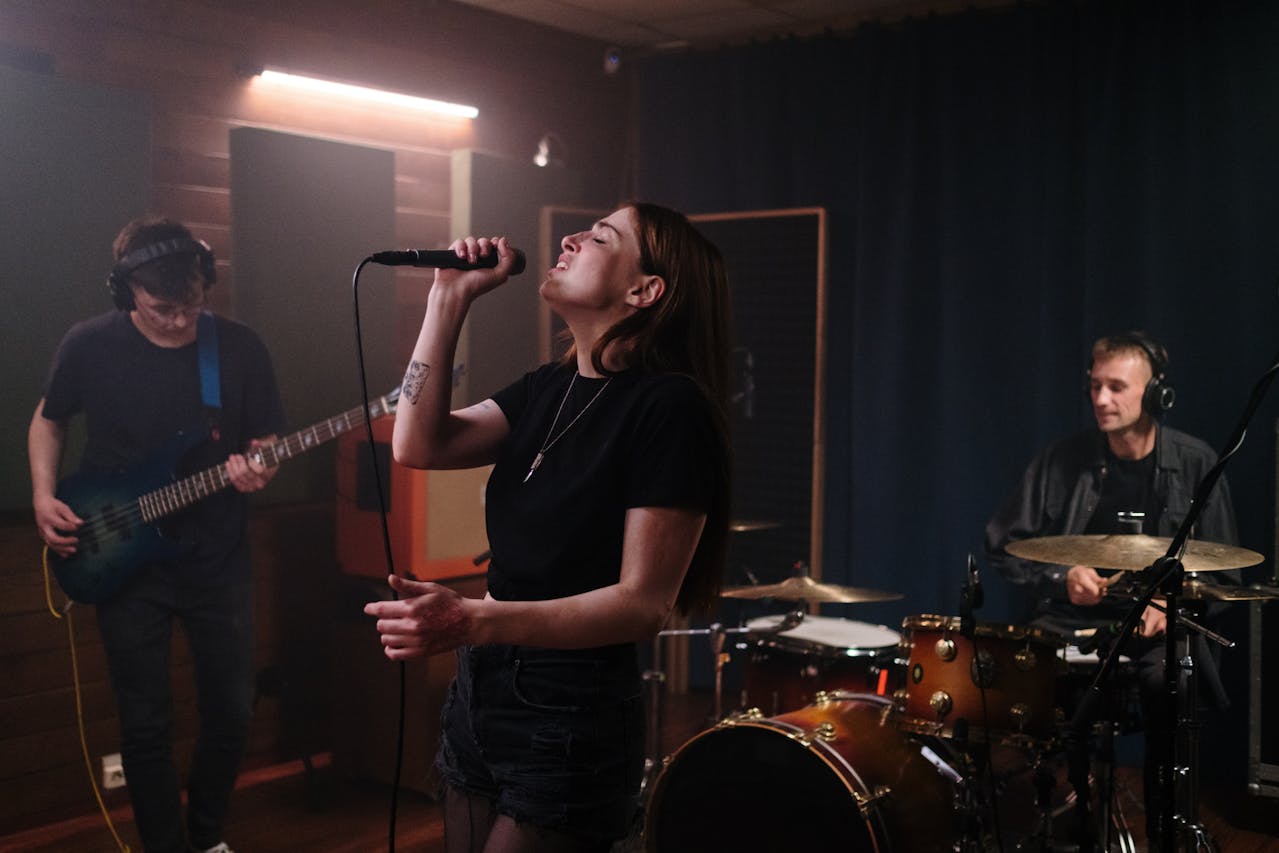 Drei Musiker proben in einem Tonstudio – eine Sängerin im Vordergrund singt leidenschaftlich ins Mikrofon, während links ein Bassist und rechts ein Schlagzeuger spielen.