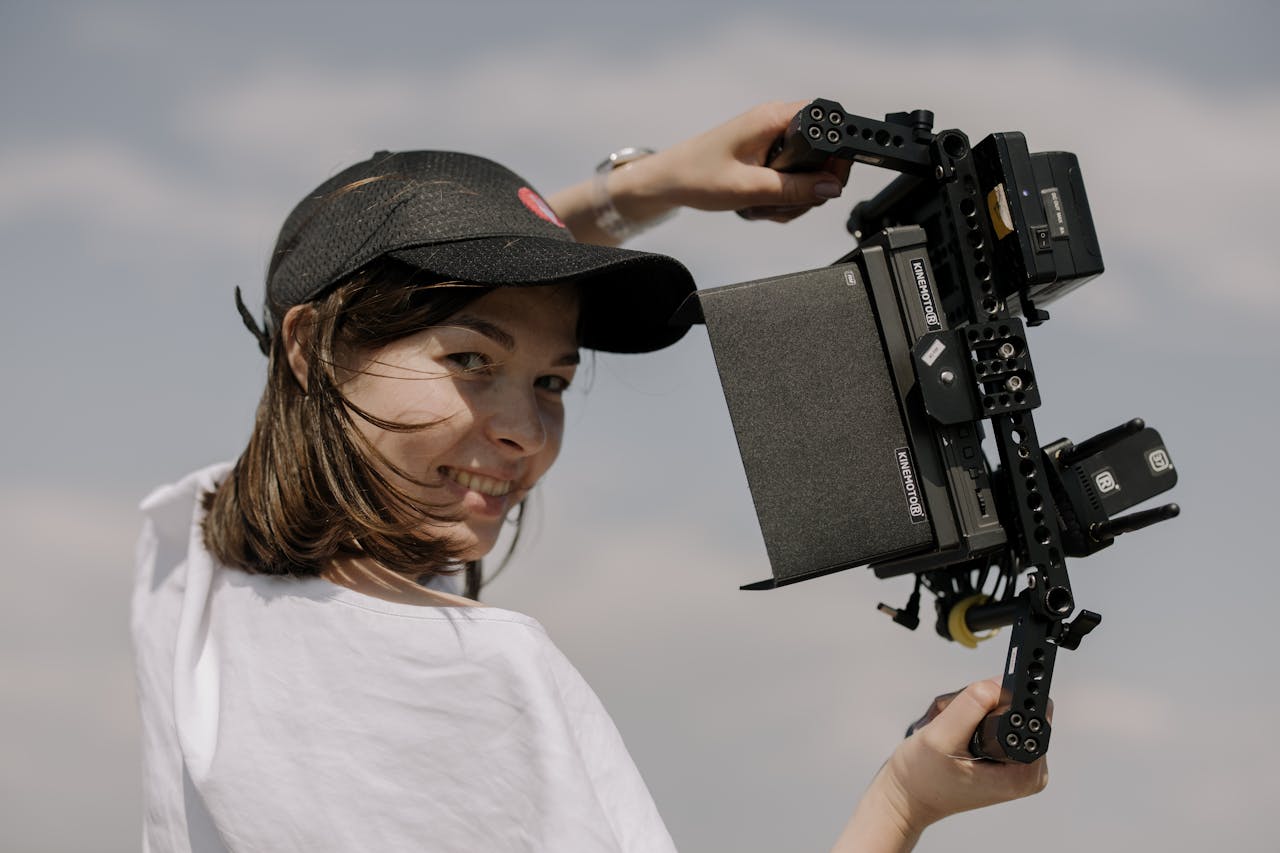 Eine junge Frau trägt ein schwarzes Basecap und hält eine große, professionelle Filmkamera über ihrer Schulter. Sie lächelt in die Kamera, während der Himmel im Hintergrund leicht bewölkt ist.