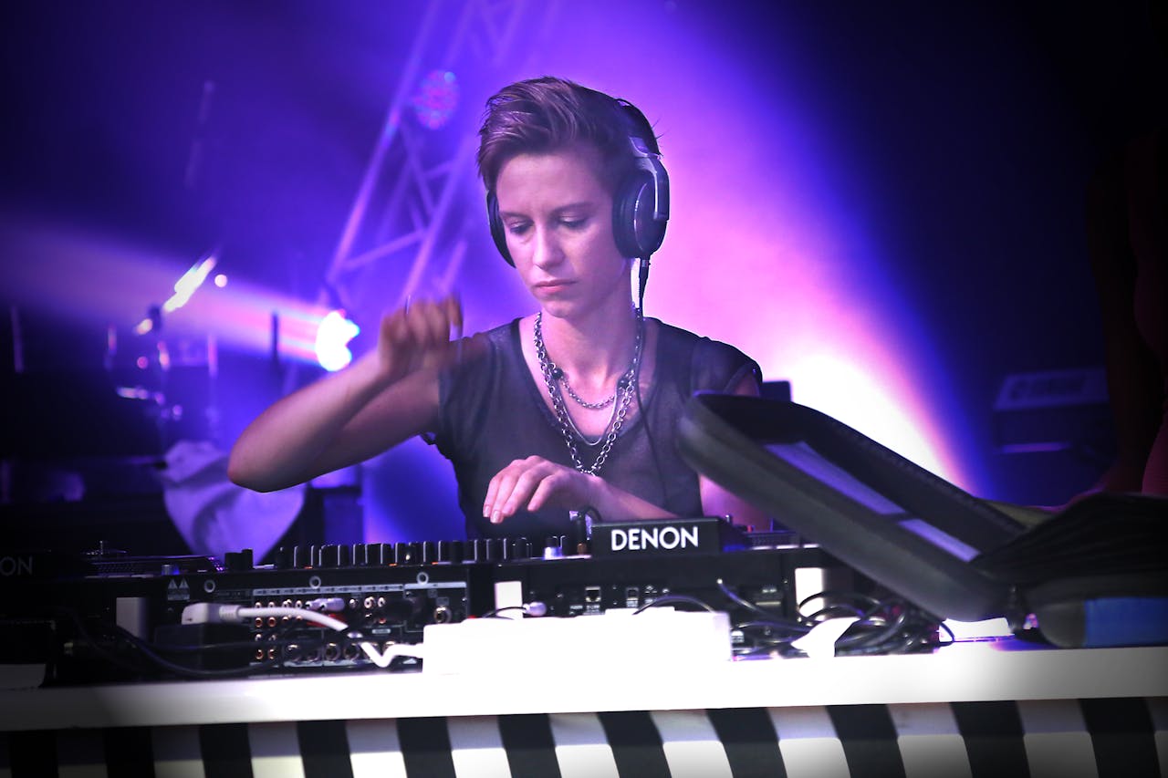 DJ mit kurzen Haaren und Kopfhörern steht an einem Mischpult der Marke Denon. Die Person trägt eine schwarze, durchsichtige Oberbekleidung und Ketten, konzentriert sich auf das Auflegen und ist von violetten Bühnenlichtern umgeben. Im Vordergrund ist ein gestreifter Tischrand sichtbar.