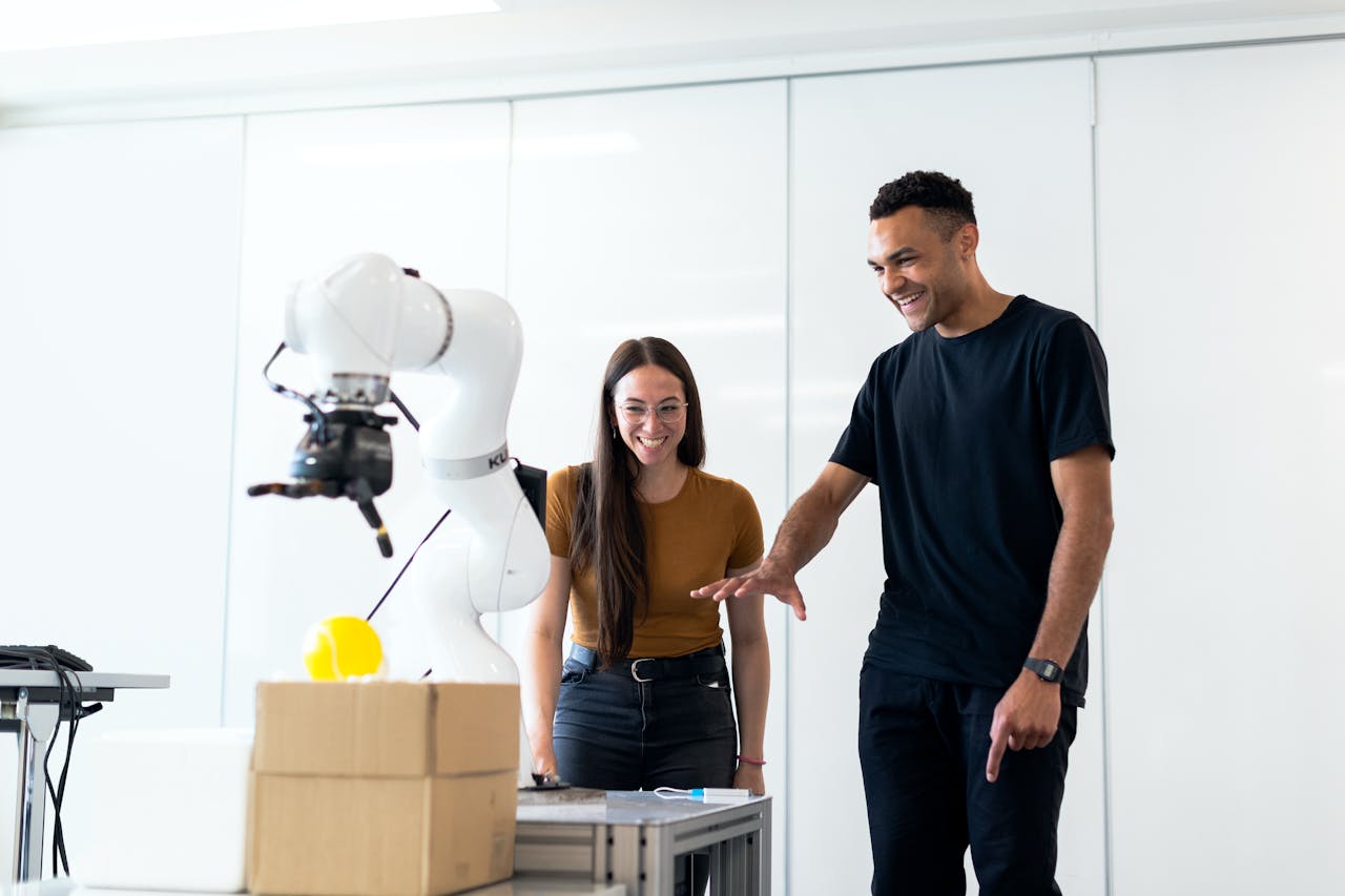 Zwei junge Erwachsene stehen lachend vor einem weißen Roboterarm in einem hellen Raum. Der Roboter bewegt einen gelben Gegenstand über einen Tisch mit einem Karton, während die beiden aufmerksam zusehen und begeistert wirken.