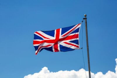 Auf dem Bild ist die Flagge des Vereinigten Königreichs (Union Jack) zu sehen, die an einem Fahnenmast im Wind weht. Der Himmel im Hintergrund ist klar blau mit einigen weißen Wolken am unteren Bildrand.