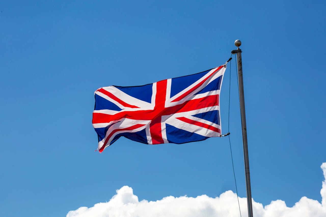 Auf dem Bild ist die Flagge des Vereinigten Königreichs (Union Jack) zu sehen, die an einem Fahnenmast im Wind weht. Der Himmel im Hintergrund ist klar blau mit einigen weißen Wolken am unteren Bildrand.