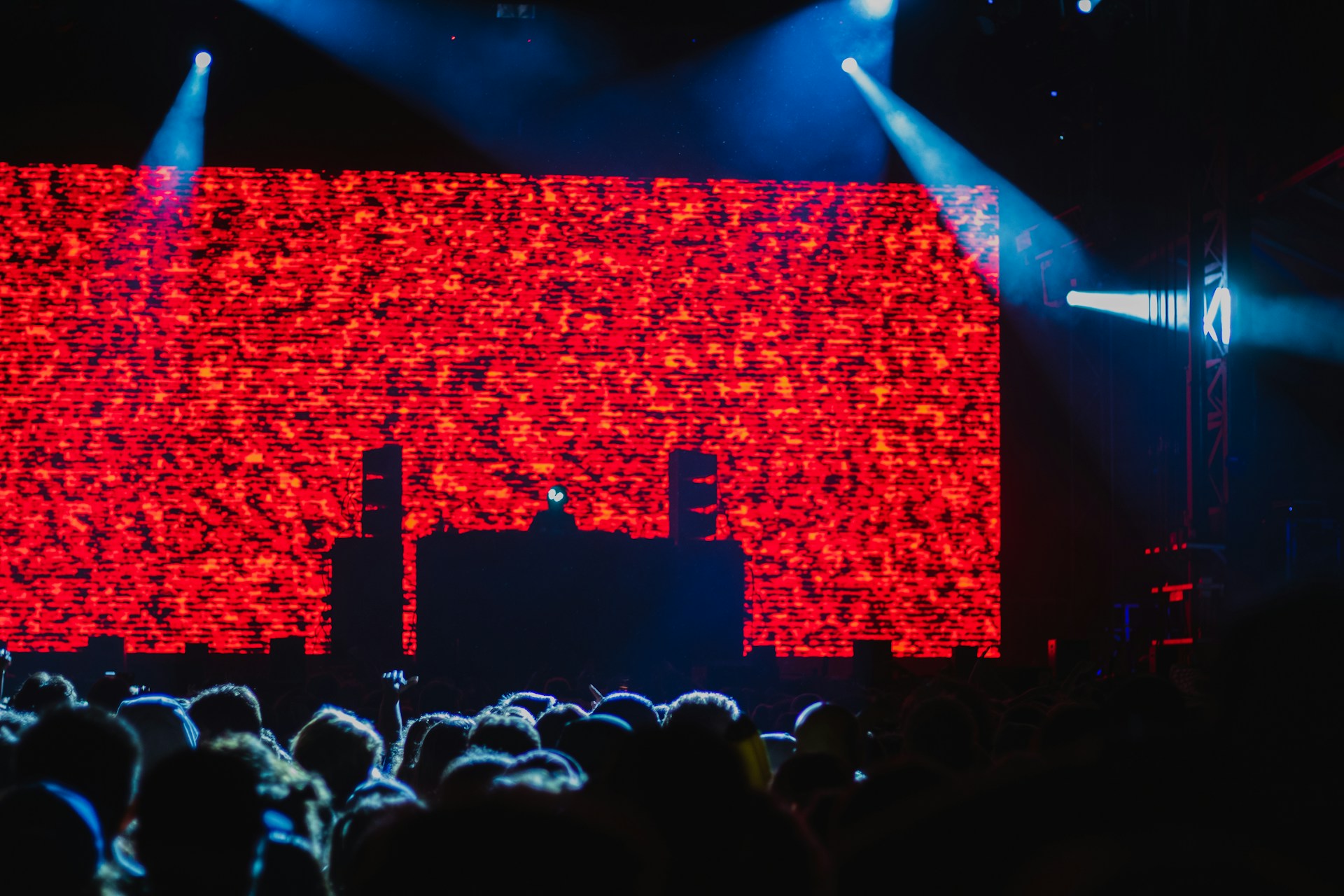 Menschenmenge auf einem Konzert oder Festival vor einer Bühne mit riesigem roten LED-Bildschirm. Silhouette eines DJs oder Künstlers am Mischpult, beleuchtet von blauen Scheinwerfern.