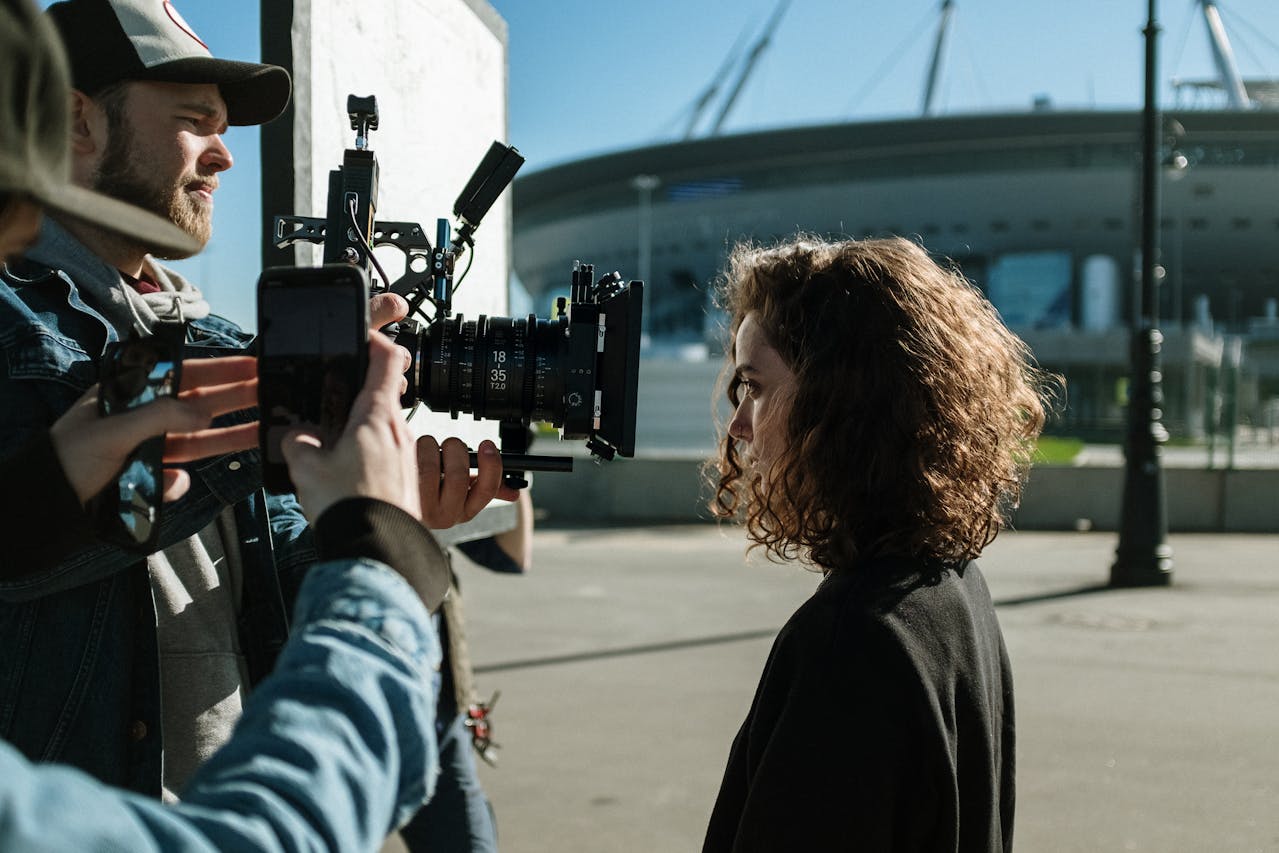 Eine Filmszene wird im Freien gedreht. Ein Kameramann mit Kappe hält eine professionelle Filmkamera auf eine Frau mit lockigen Haaren, die im Profil zu sehen ist. Neben der Kamera ist eine weitere Person mit Smartphone sichtbar. Im Hintergrund steht ein modernes Stadion.