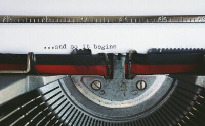 Nahaufnahme einer Schreibmaschine, die auf ein Blatt Papier den Satz „...and so it begins“ tippt.