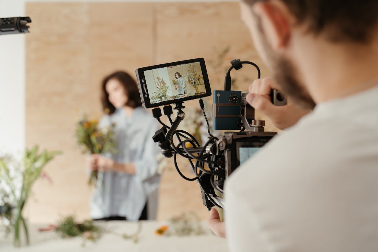 Eine Person filmt mit einer professionellen Kamera eine Frau, die in einem hellen Raum Blumenarrangements hält. Auf dem Kamerabildschirm ist die Frau scharf zu sehen, während sie im Hintergrund unscharf bleibt.