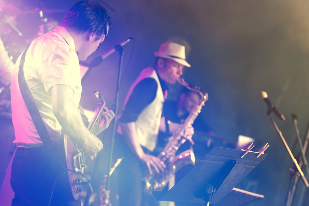Zwei Musiker auf einer Bühne in atmosphärischer blau-violetter Beleuchtung mit Nebel-Effekten. Im Vordergrund spielt ein Gitarrist im weißen Hemd eine E-Gitarre, während im Hintergrund ein Saxophonist mit Fedora-Hut musiziert. Unten rechts ist ein Notenständer mit Notenblättern sichtbar.