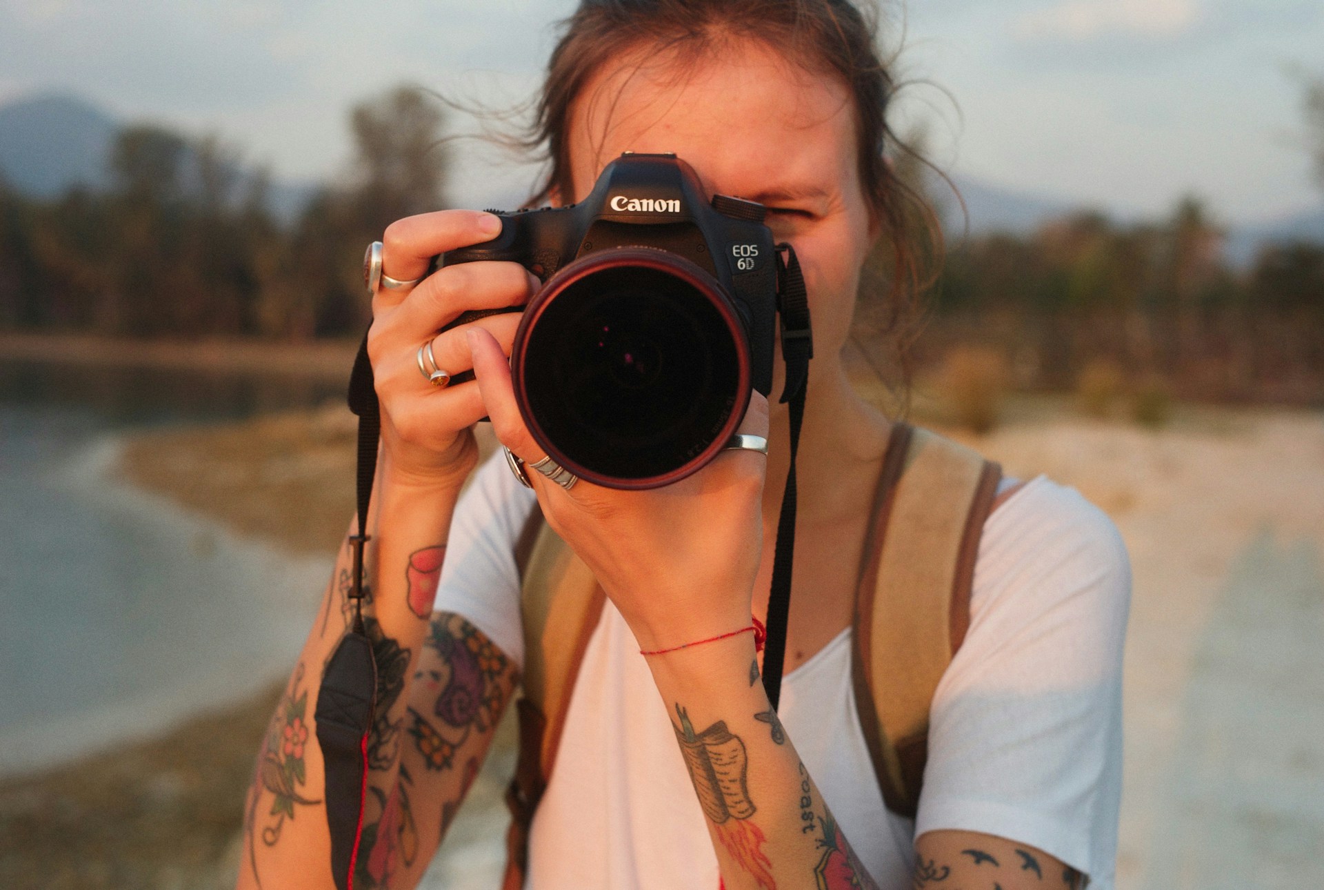 Person mit tätowierten Armen und weißem T-Shirt hält eine Canon-Kamera vor das Gesicht und macht ein Foto im Freien, mit unscharfem Hintergrund aus Wasser, Bäumen und Bergen bei warmem Licht.