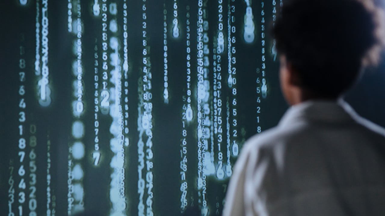Eine Person steht vor einem großen Bildschirm oder einer Projektion, auf der in grünlich-weißer Farbe Zahlen vertikal von oben nach unten laufen. Die Szene erinnert an digitale Codes oder Datenströme und vermittelt eine technologische, futuristische Atmosphäre. Die Person ist von hinten zu sehen und trägt ein helles Oberteil.