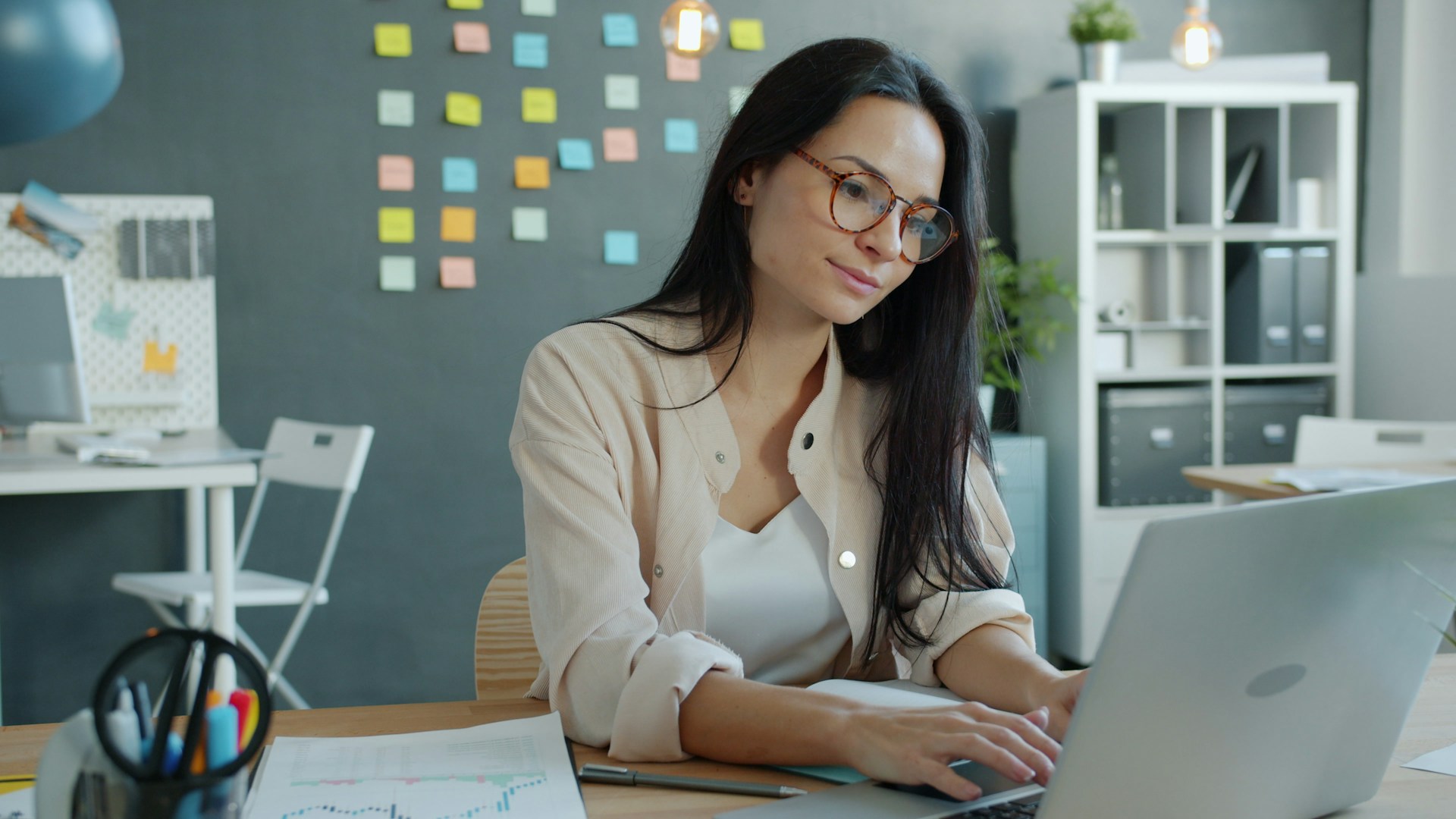 Die Szene zeigt eine Person, die konzentriert an einem Laptop arbeitet. Sie sitzt an einem ordentlich eingerichteten Schreibtisch, auf dem einige Unterlagen mit Diagrammen sowie Büro-Utensilien liegen. Im Hintergrund hängen bunte Haftnotizen an einer dunklen Wand, was nach einem kreativen oder projektorientierten Arbeitsumfeld aussieht. Regale mit Ordnern, Pflanzen und Schreibtischlampen ergänzen den Raum und verleihen ihm eine moderne, strukturierte Atmosphäre. Insgesamt wirkt das Bild wie eine ruhige, produktive Bürosituation – jemand, der fokussiert an einer Aufgabe oder einem Projekt sitzt.