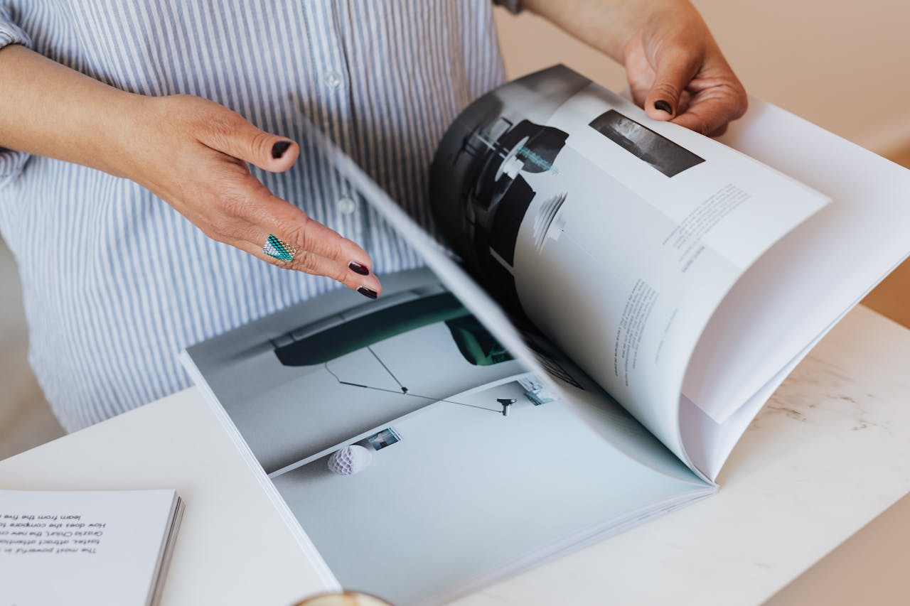 Eine Person mit dunklem Nagellack und türkisfarbenem Ring blättert in einem großformatigen Buch oder Katalog, der auf einem weißen Tisch liegt. Die Seiten zeigen Fotos moderner Raumgestaltung in minimalistischen Farben. Die Person trägt ein hellblau-weiß gestreiftes Hemd. Die Szene wirkt ruhig und konzentriert, mit Fokus auf die Hände und das Buch.