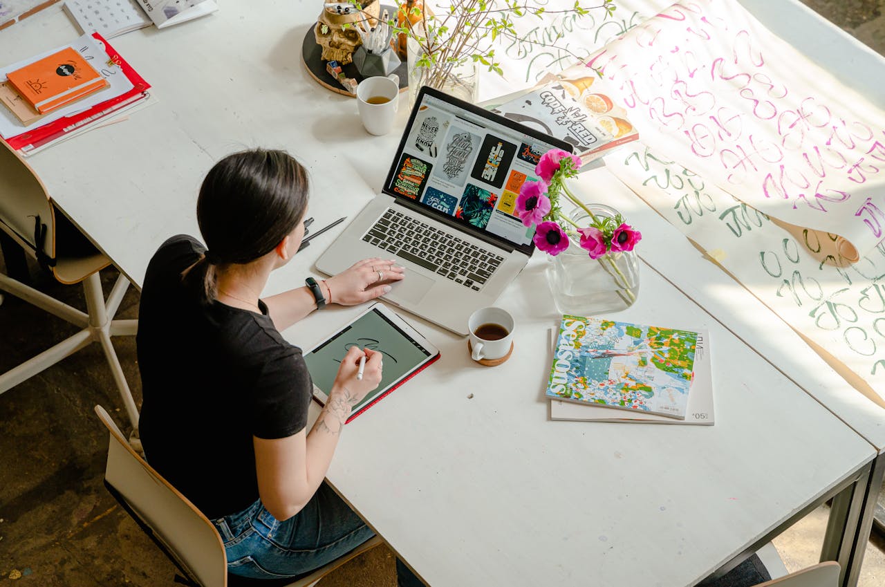 Hier ist ein Alt-Text für das Bild: "Vogelperspektive auf eine kreative Designerin an einem großen weißen Arbeitstisch. Sie arbeitet mit einem Stylus auf einem Grafiktablet, während vor ihr ein Laptop mit Designentwürfen geöffnet ist. Auf dem Tisch befinden sich eine Tasse Tee, pinke Mohnblumen in einer Vase, Magazine, Notizbücher und große Papierbögen mit handgeschriebenen Schriftzügen in verschiedenen Farben. Die Szene zeigt einen typischen kreativen Arbeitsplatz.