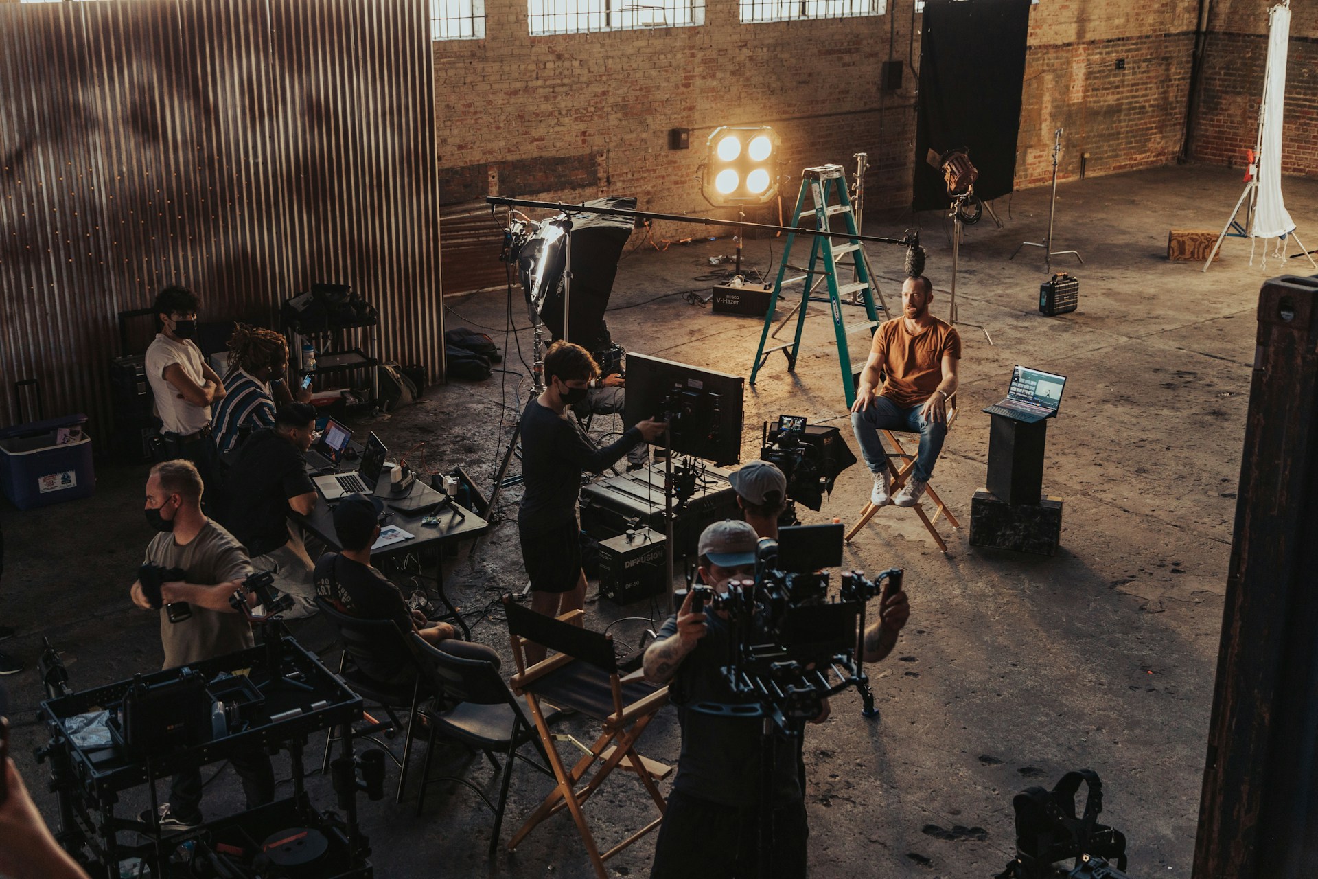 "Filmcrew bei Dreharbeiten in einer industriellen Lagerhalle mit Backsteinwänden. Ein Kameramann filmt eine Person, die auf einem Regiestuhl sitzt, während Crewmitglieder verschiedene Ausrüstung bedienen, darunter Monitore, Scheinwerfer und eine Leiter. Professionelles Videoproduktions-Setup mit mehreren koordiniert arbeitenden Teammitgliedern