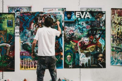 Ein Künstler in weißem T-Shirt und farbverschmierter Hose steht von hinten fotografiert vor einer weißen Wand, an der mehrere großformatige Collagen und Mixed-Media-Kunstwerke hängen. Er arbeitet gerade an einem der bunten, layerten Werke, die Textelemente wie 'EVA', 'AVEC LE CŒUR' und verschiedene Graffiti-Stile, Porträts und abstrakte Elemente in leuchtenden Farben – vor allem Blau-, Grün- und Rottönen – kombinieren. Seine Kleidung zeigt Farbspuren seiner künstlerischen Arbeit.