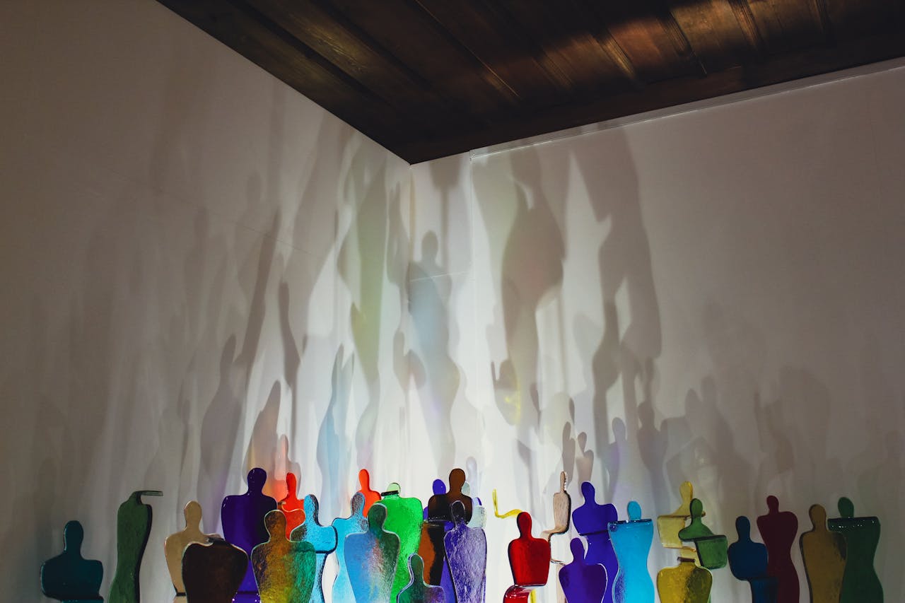Kunstinstallation in einem Raum mit dunkler Holzdecke: Eine Reihe bunter, durchscheinender Glasfiguren in Menschenform steht vor einer weißen Wand. Die Figuren in verschiedenen Farben – Blau, Grün, Rot, Gelb, Orange und Lila – werden von hinten beleuchtet, wodurch ihre vergrößerten, verschwommenen Schatten an die Wand projiziert werden. Die Schatten überlagern sich und erzeugen ein ätherisches, traumhaftes Schattenspiel.