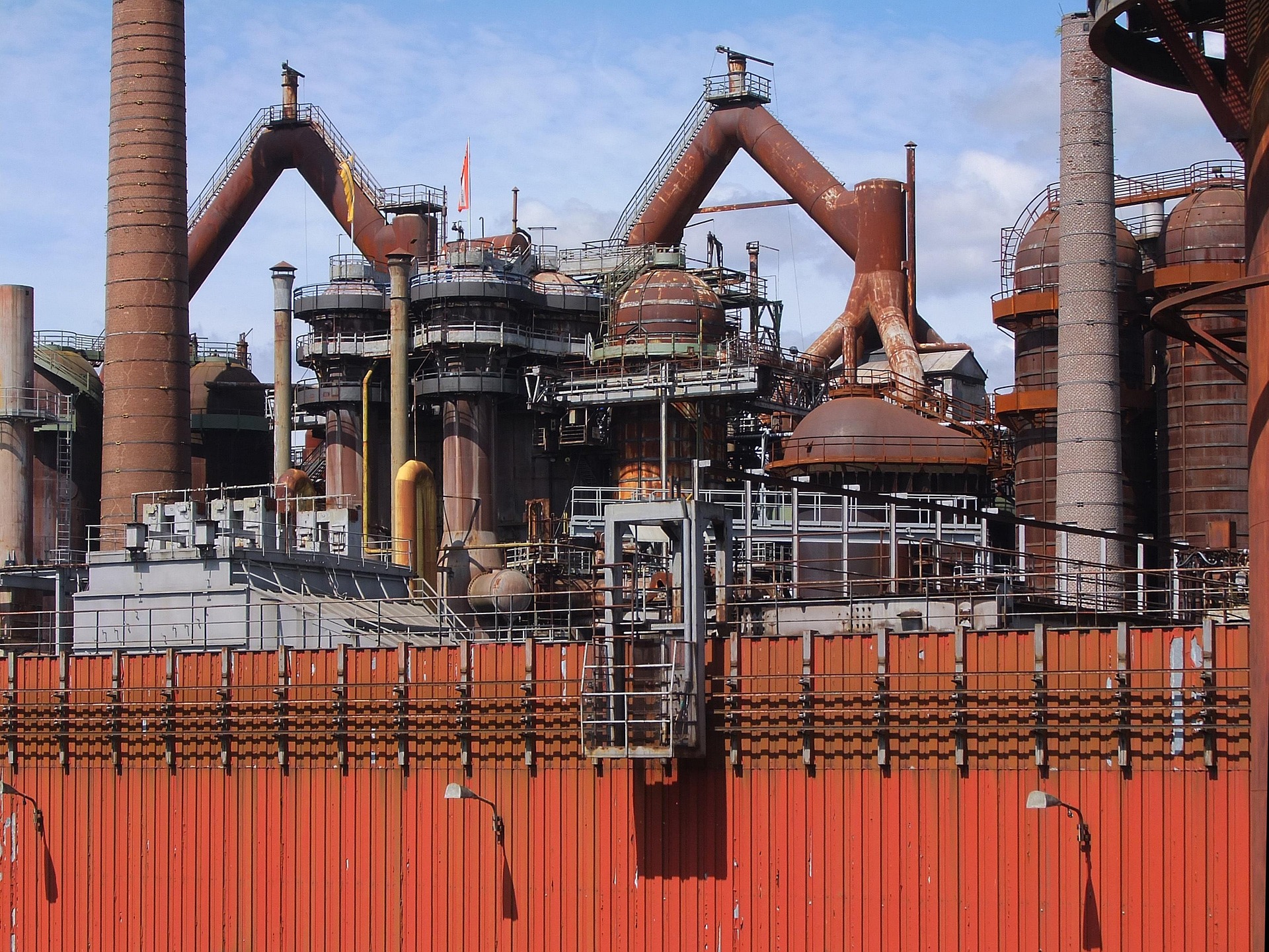 Historisches Industriedenkmal einer stillgelegten Hüttenanlage mit charakteristischen rostbraunen Hochöfen, Schornsteinen und gebogenen Gasleitungen. Im Vordergrund eine orange-rote Wellblechzaunwand, dahinter das komplexe System aus Rohrleitungen, Treppen und Plattformen der ehemaligen Eisenproduktionsanlage vor blauem Himmel.