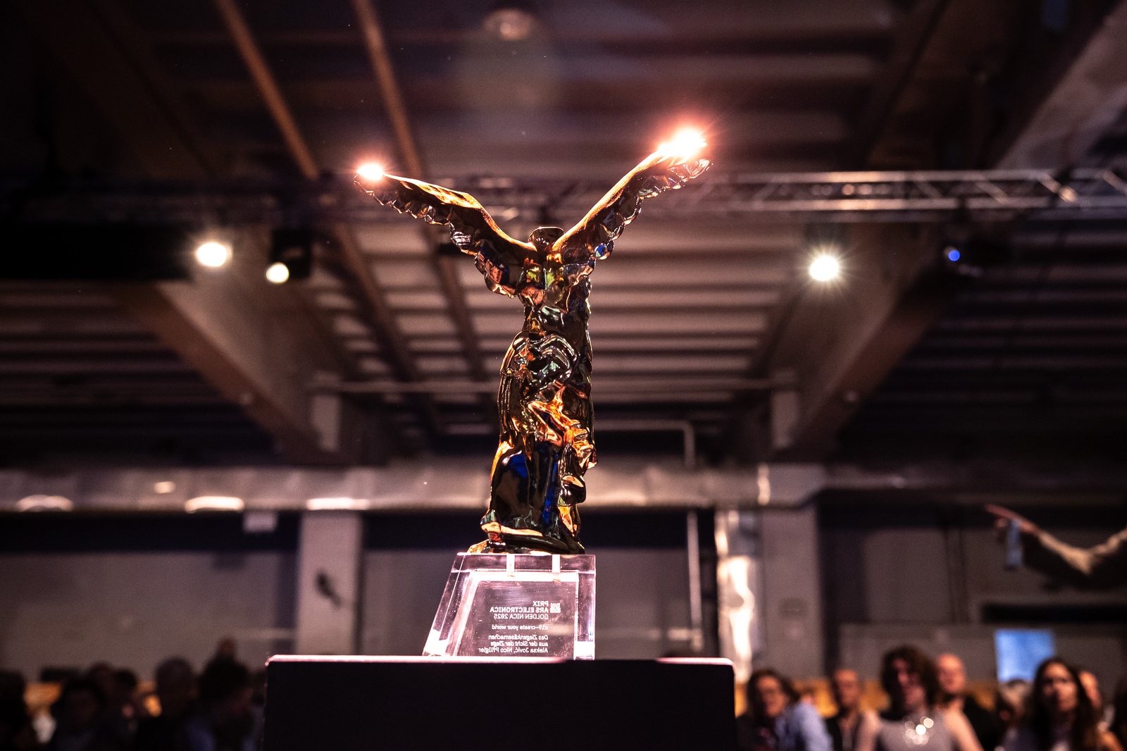 Eine goldene Trophäe in Form einer Siegesgöttin mit ausgebreiteten Flügeln steht auf einem transparenten Kristallsockel. Die Statue wird dramatisch von oben beleuchtet, wodurch die Flügelspitzen hell erstrahlen. Im unscharfen Hintergrund ist eine Veranstaltungshalle mit Industriedecke und Publikum zu erkennen. Die Atmosphäre wirkt festlich und feierlich, wie bei einer Preisverleihung.