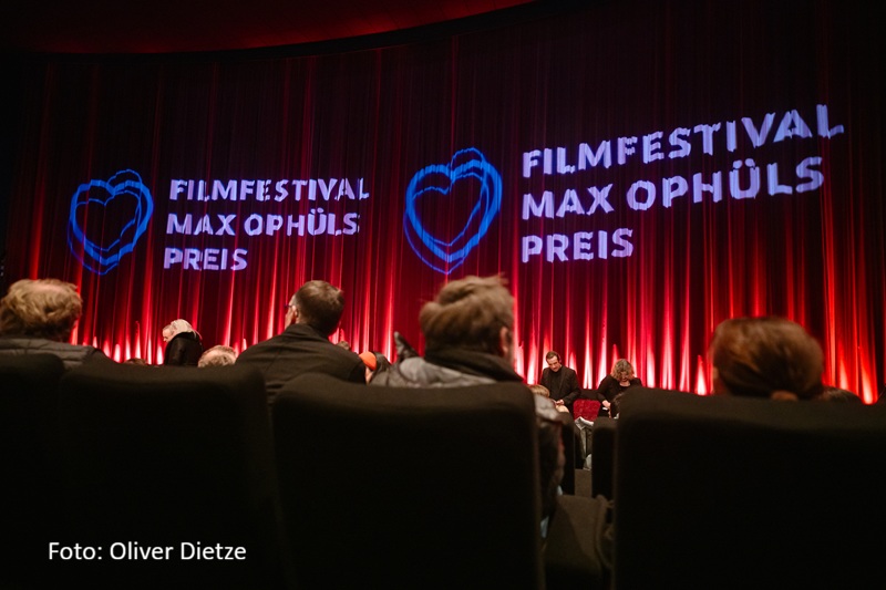 Blick aus dem Zuschauerraum auf eine Kinoleinwand mit rot beleuchtetem Vorhang. Auf der Leinwand ist zweimal das Logo des Filmfestivals Max Ophüls Preis in blau-violetter Schrift projiziert, jeweils begleitet von dem charakteristischen Herz-Symbol aus drei verschlungenen Herzlinien. Im Vordergrund sieht man die Silhouetten der sitzenden Zuschauer, während auf der Bühne zwei Personen zu erkennen sind.
