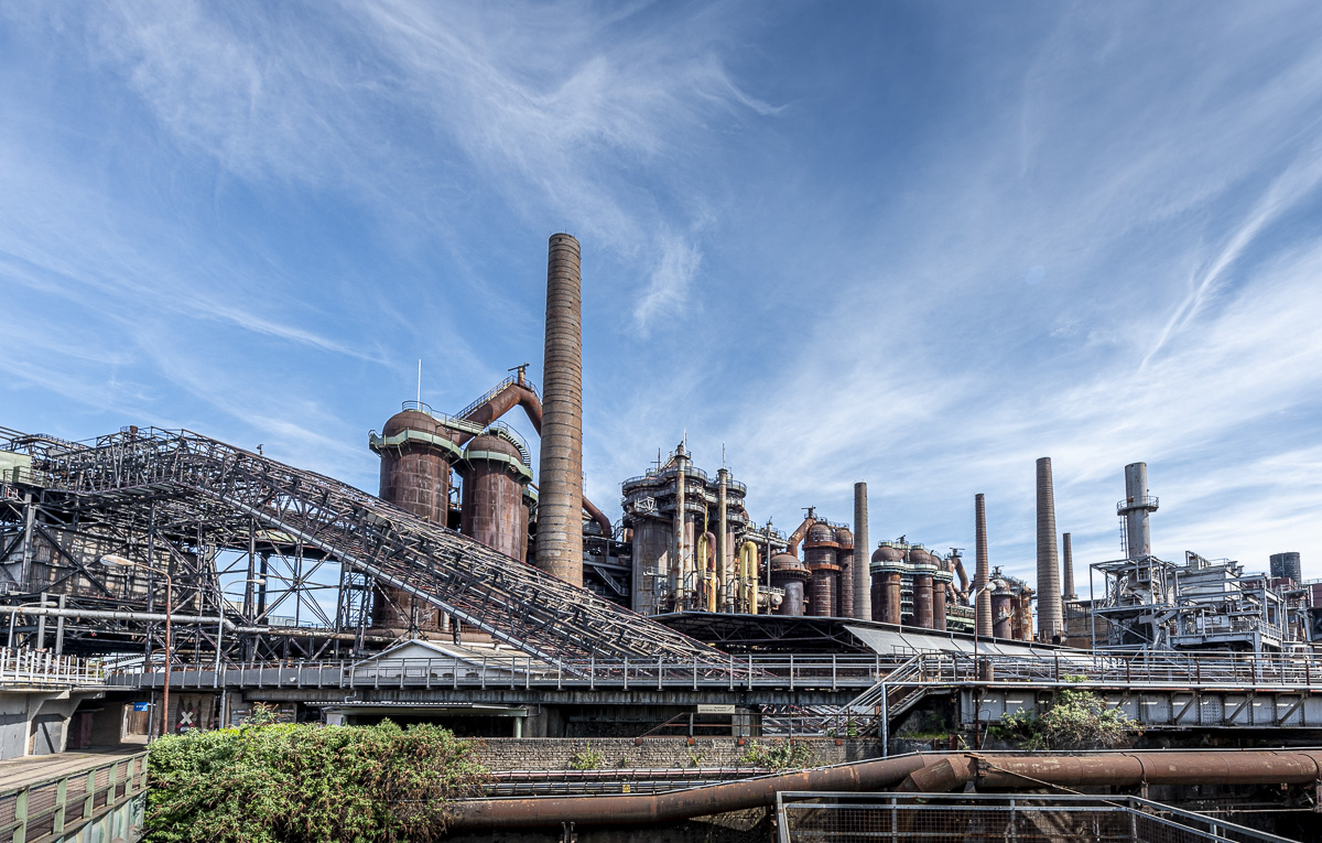 Panoramaansicht der Völklinger Hütte, einem UNESCO-Weltkulturerbe. Die monumentale Industrieanlage zeigt massive Hochöfen in Rostbraun, hohe Schornsteine, metallene Förderbänder und ein komplexes System aus Stahlkonstruktionen, Rohrleitungen und Plattformen. Im Vordergrund grüne Vegetation, darüber ein dramatischer blauer Himmel mit Zirruswolken.