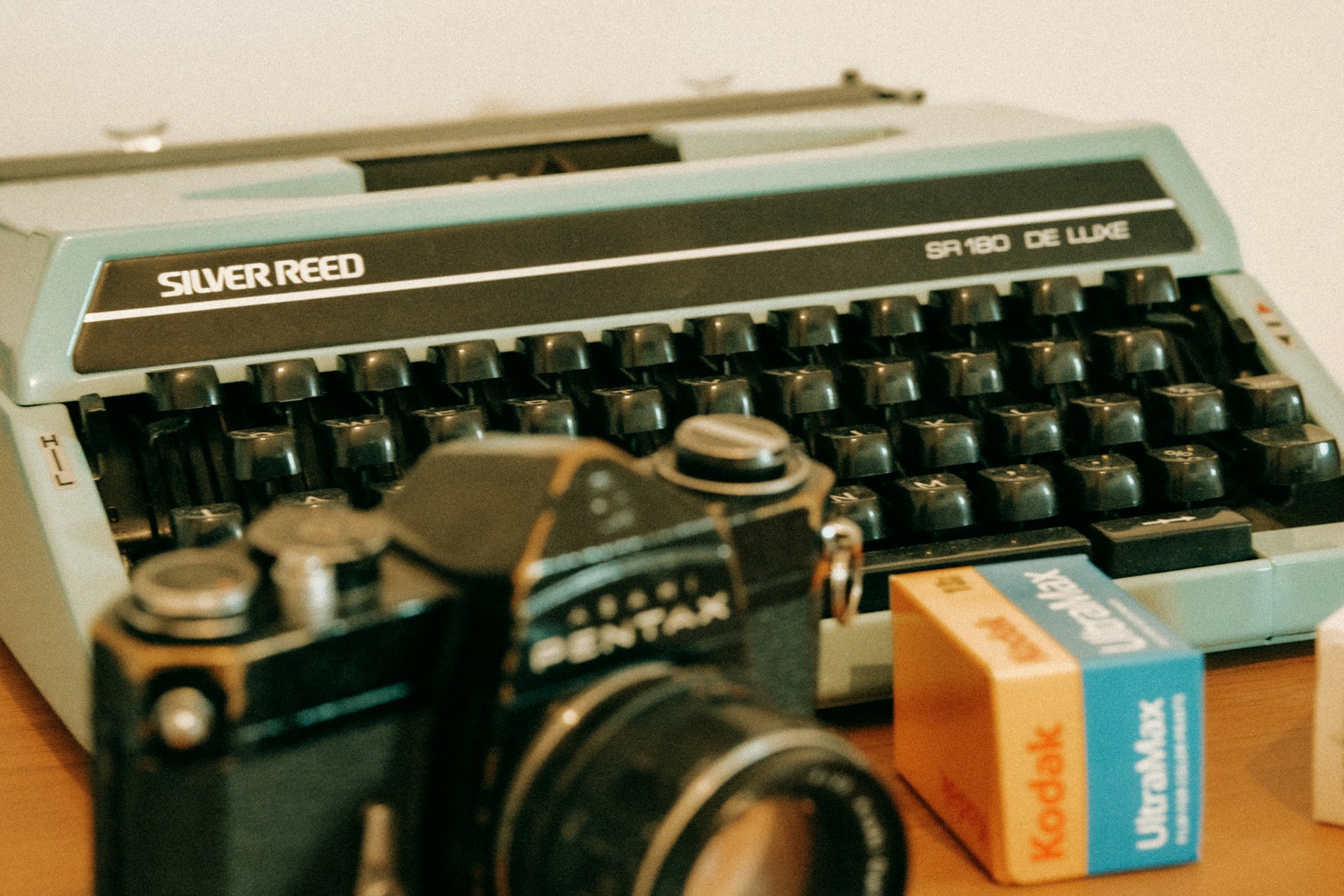 Nostalgische Nahaufnahme einer vintage Silver Reed SR 180 De Luxe Schreibmaschine in Hellblau mit schwarzen Tasten. Im Vordergrund liegen eine analoge Filmkamera und Kodak Ultramax Kleinbildfilme in oranger und blauer Verpackung. Die Komposition vermittelt eine warme, vintage Ästhetik der analogen Ära