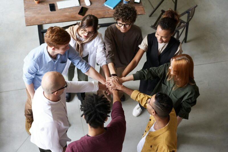 Vogelperspektive auf eine diverse Gruppe von acht Personen, die ihre Hände in der Mitte eines Kreises zusammenlegen – eine Geste des Teamgeists und der Zusammenarbeit. Das Team steht in einem modernen Büroraum mit hellem Boden, im Hintergrund ein Holztisch mit Computern, Notizbüchern und Smartphones. Die Teammitglieder tragen lässige Business-Kleidung in verschiedenen Farben.